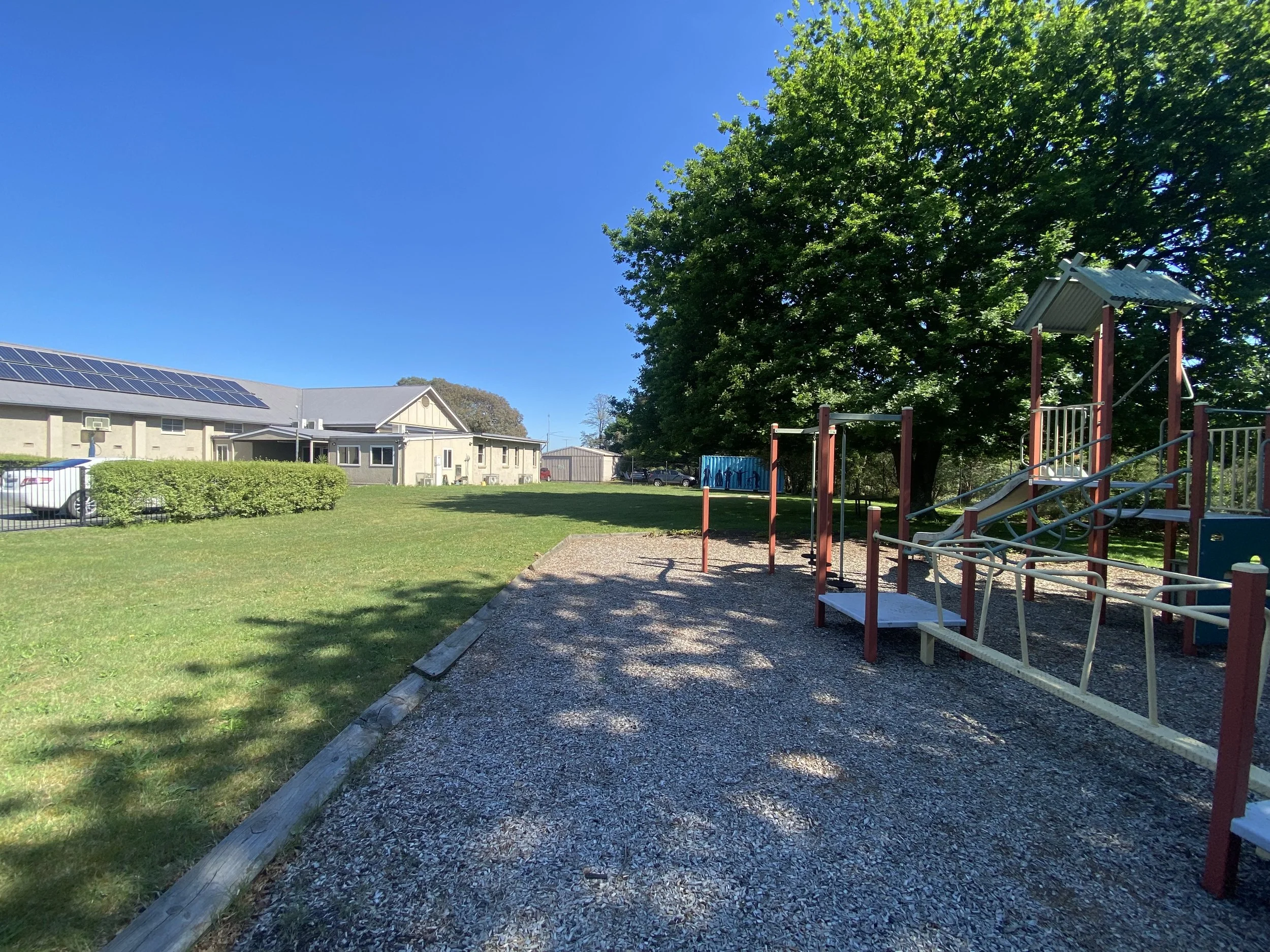 A playground with a slide, climbing bars, and swings situated on gravel, with a large tree and a building with solar panels in the background on a clear, sunny day.