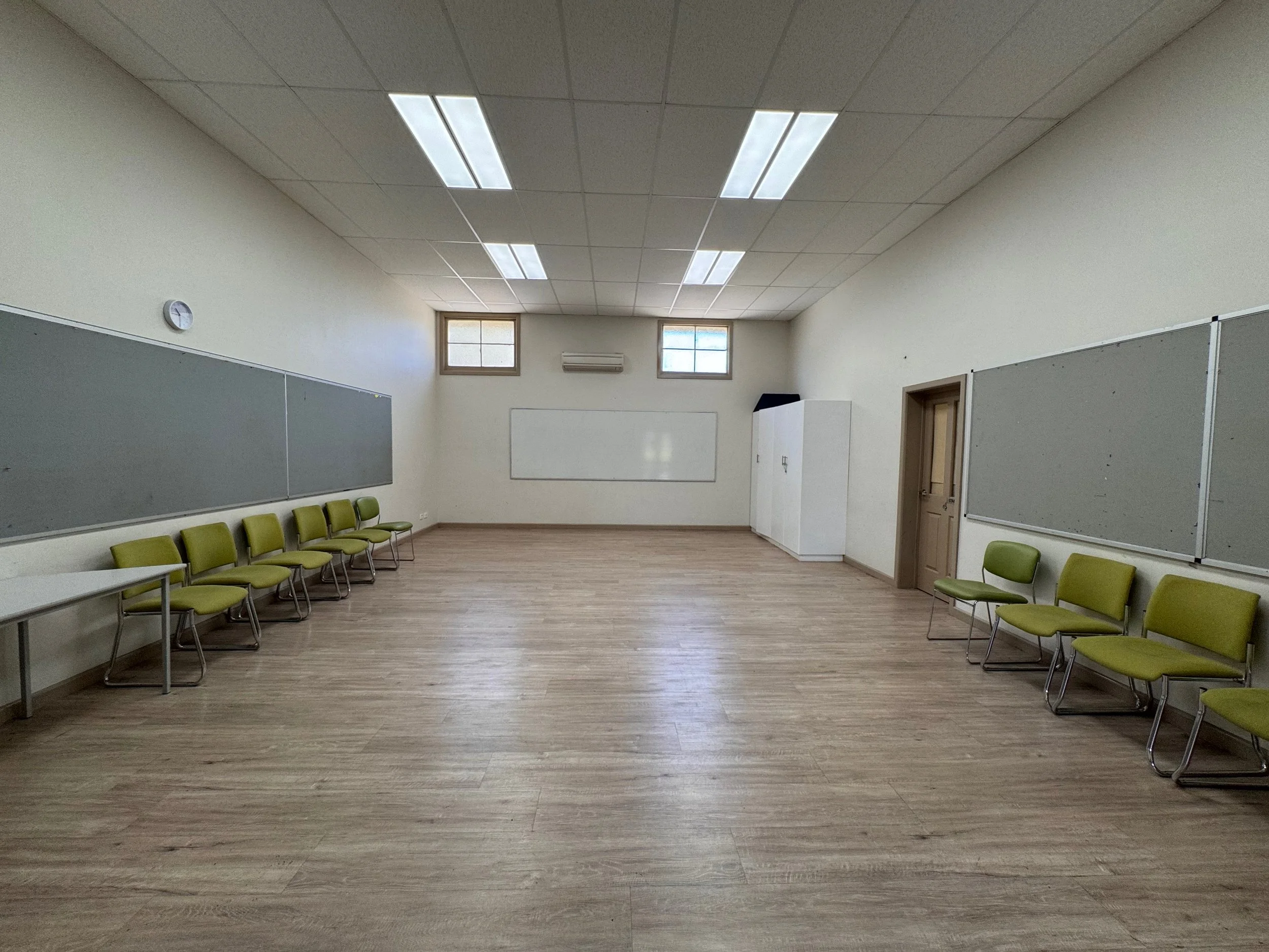 Empty classroom with wooden floor, green chairs arranged along the walls, large blank whiteboards, small wall clock, ceiling lights, and a window.
