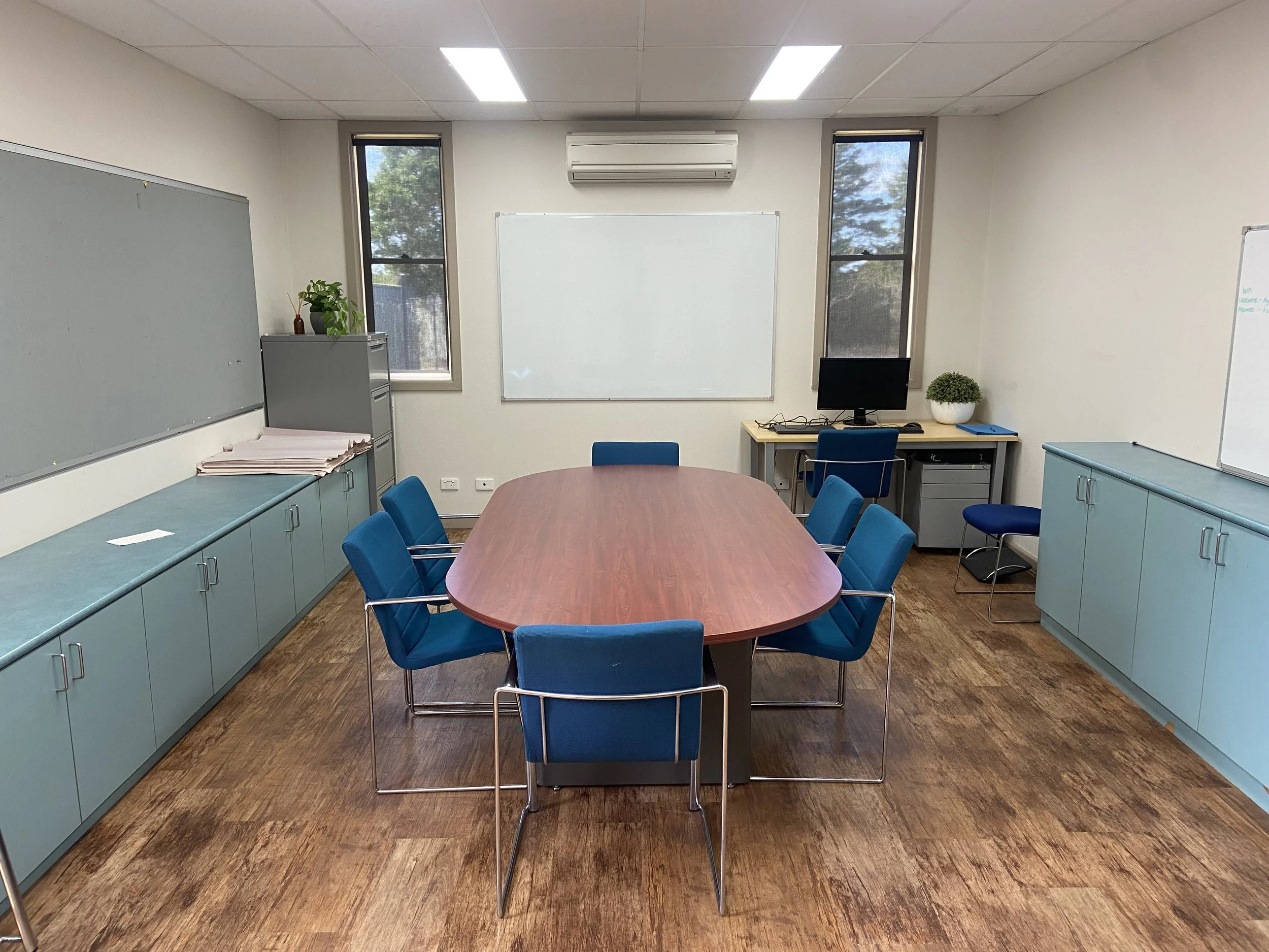 Conference room with oval table, blue chairs, computer desk, and cabinets. Whiteboard and air conditioning unit on walls.