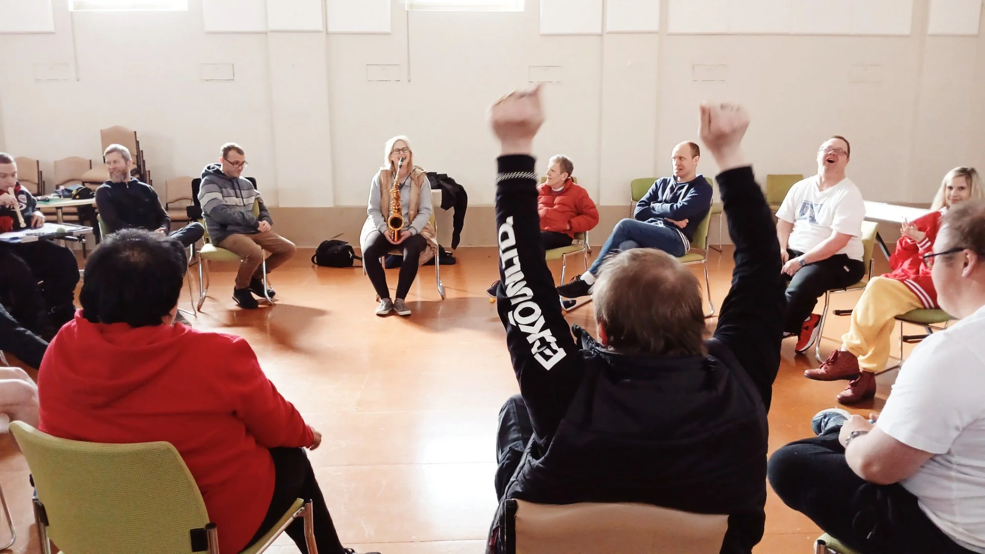 A group of people sitting in a circle in a large room, while a woman in the center plays a saxophone. The atmosphere appears relaxed and some participants look engaged or cheerful.