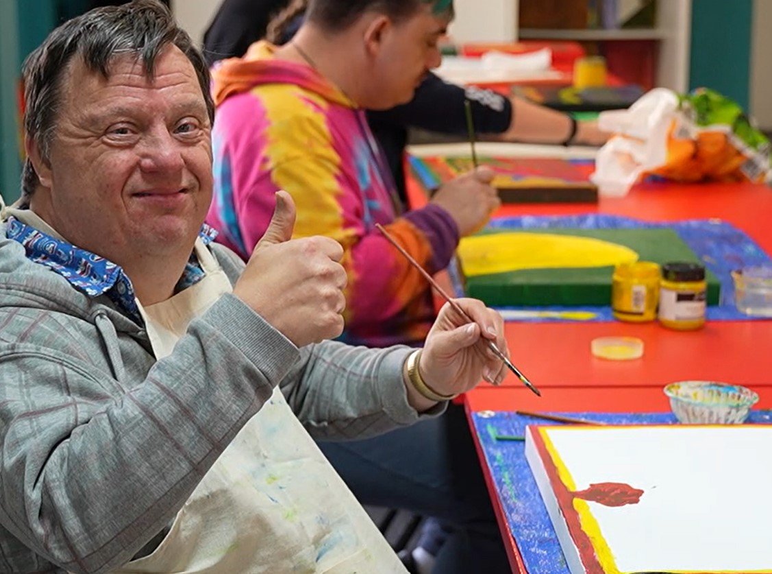 Man smiling and giving a thumbs-up while painting, seated at a table with art supplies.