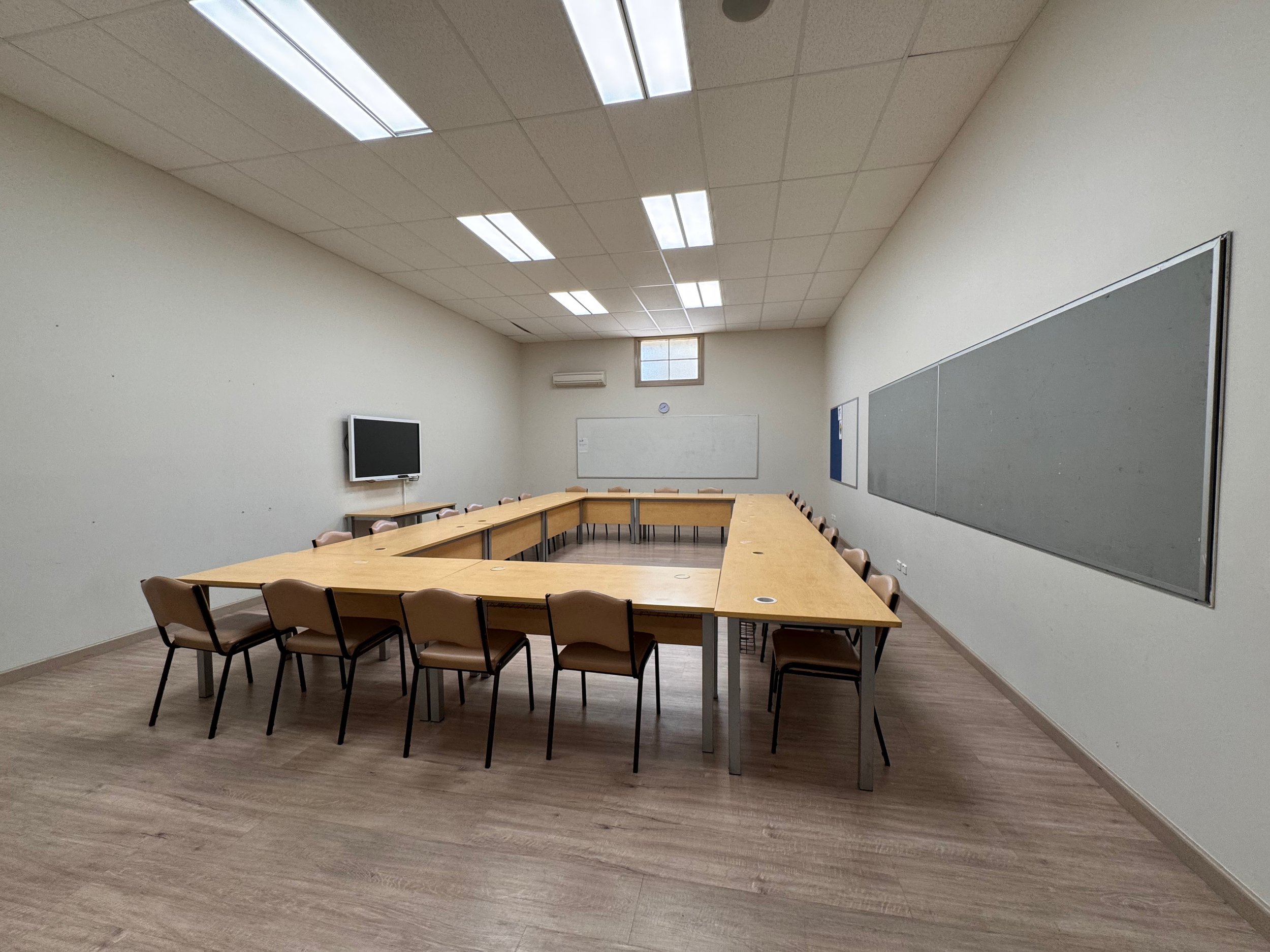 Empty conference or classroom with U-shaped table, chairs, whiteboard, and wall-mounted monitor.