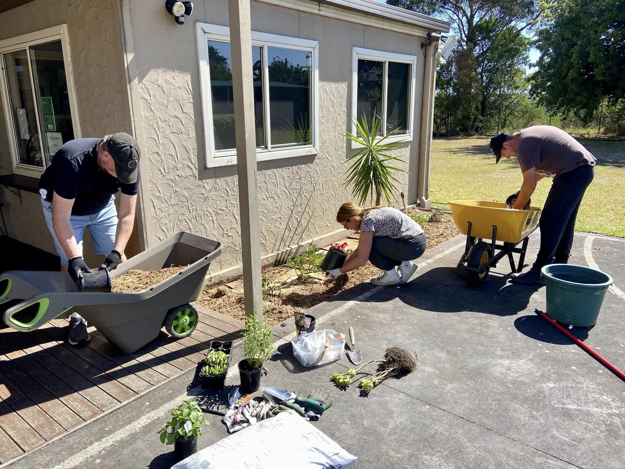 Three people working together outdoors, planting flowers and doing yard work near a house on a sunny day.