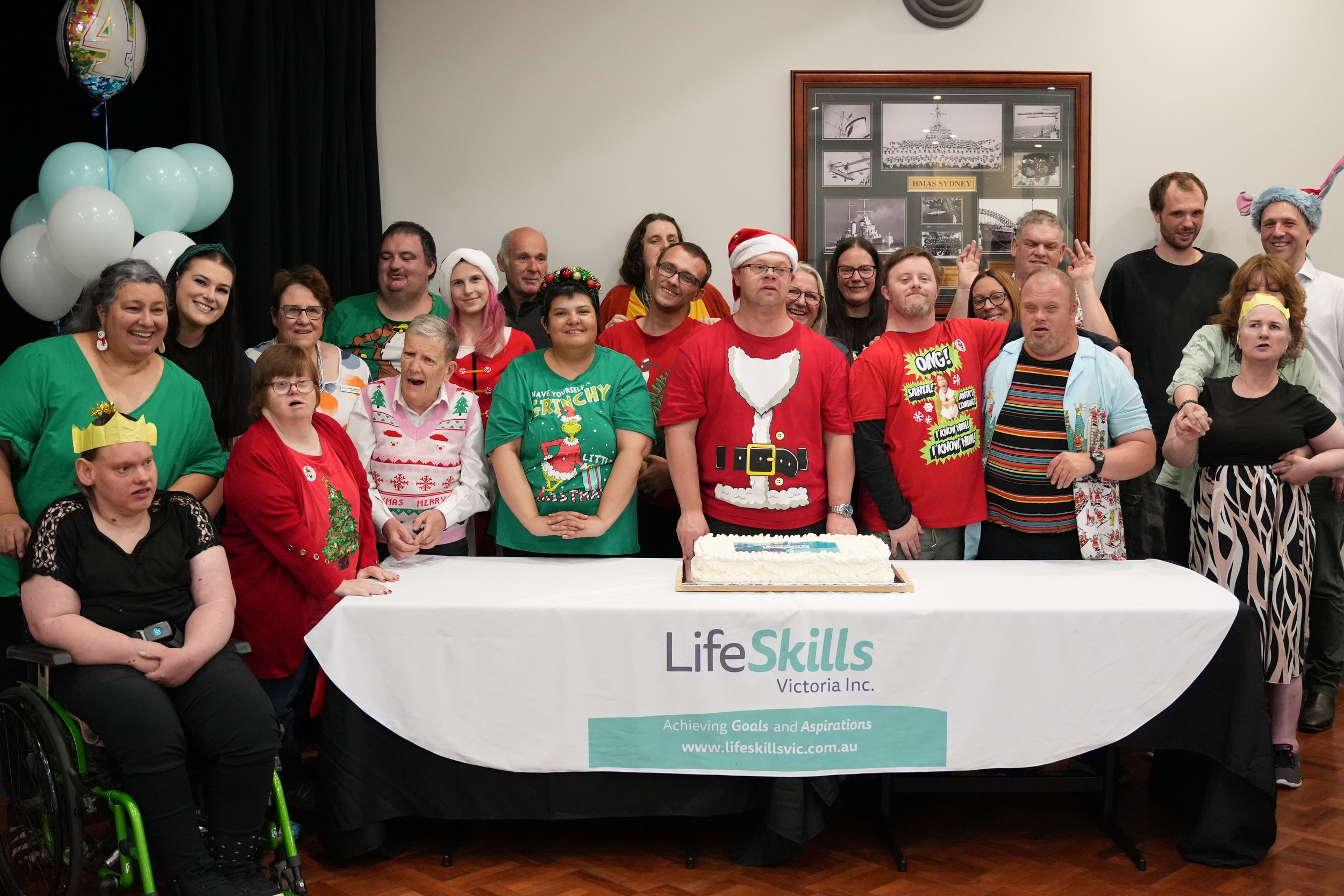 A group of people at a festive gathering, some wearing holiday-themed clothing, standing behind a table with a cake. The tablecloth displays the logo and name "LifeSkills Victoria Inc." along with their slogan and website. Balloons are visible in the background.