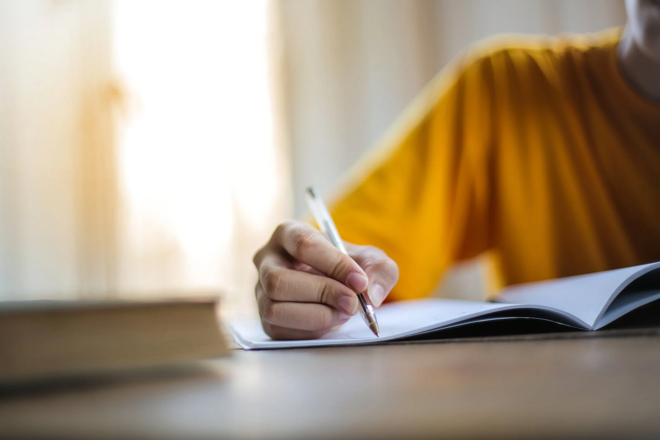 Person writing in a notebook with a pen, wearing a yellow shirt, indoors.