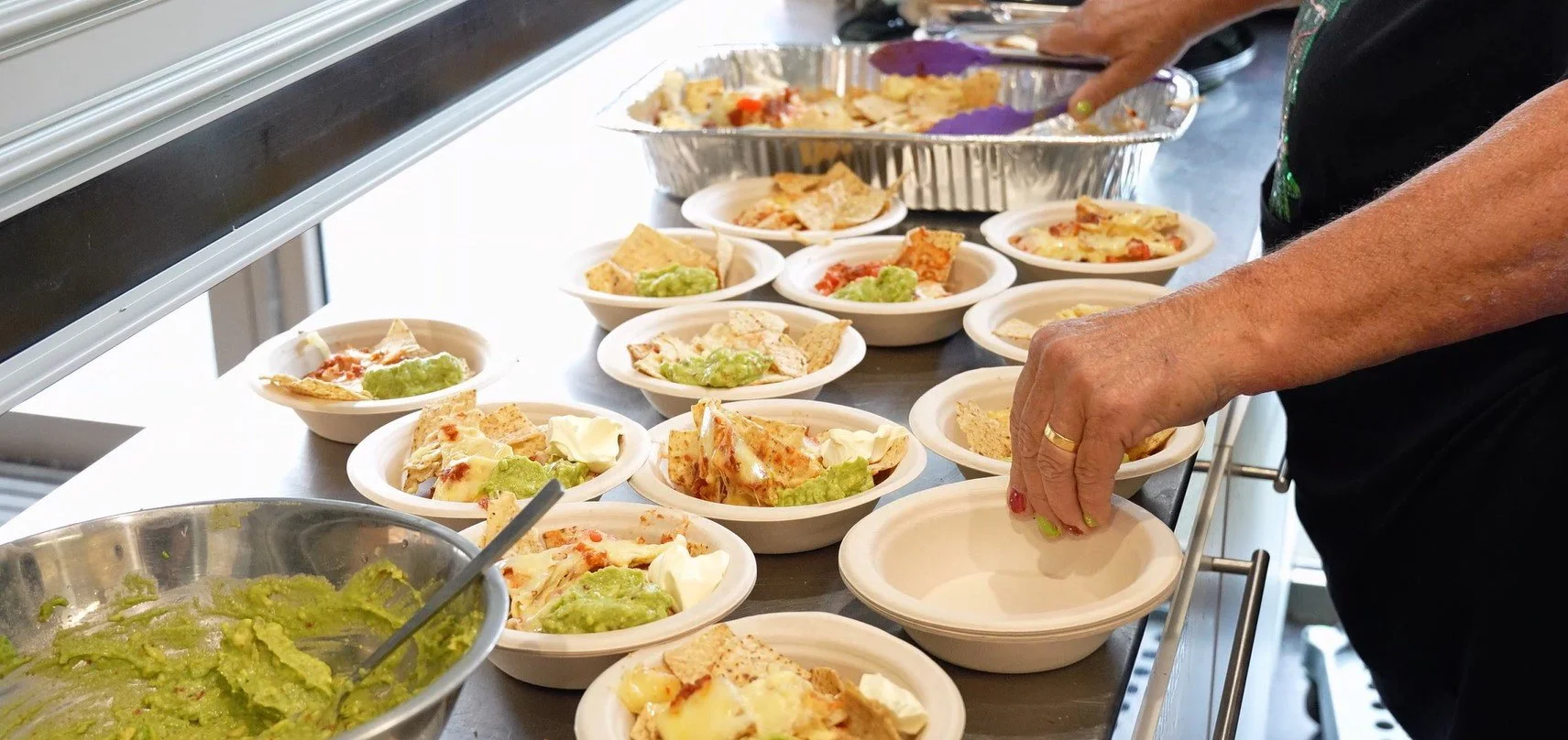 Person preparing multiple plates of nachos with guacamole, cheese, and salsa.