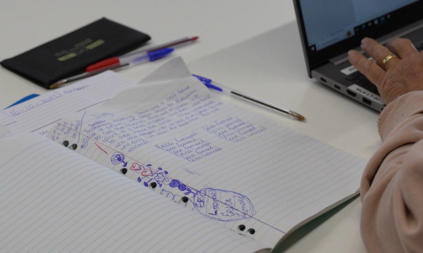 Open notebook with handwritten notes and drawings, pens, a person typing on a laptop, and a small black case on a desk.