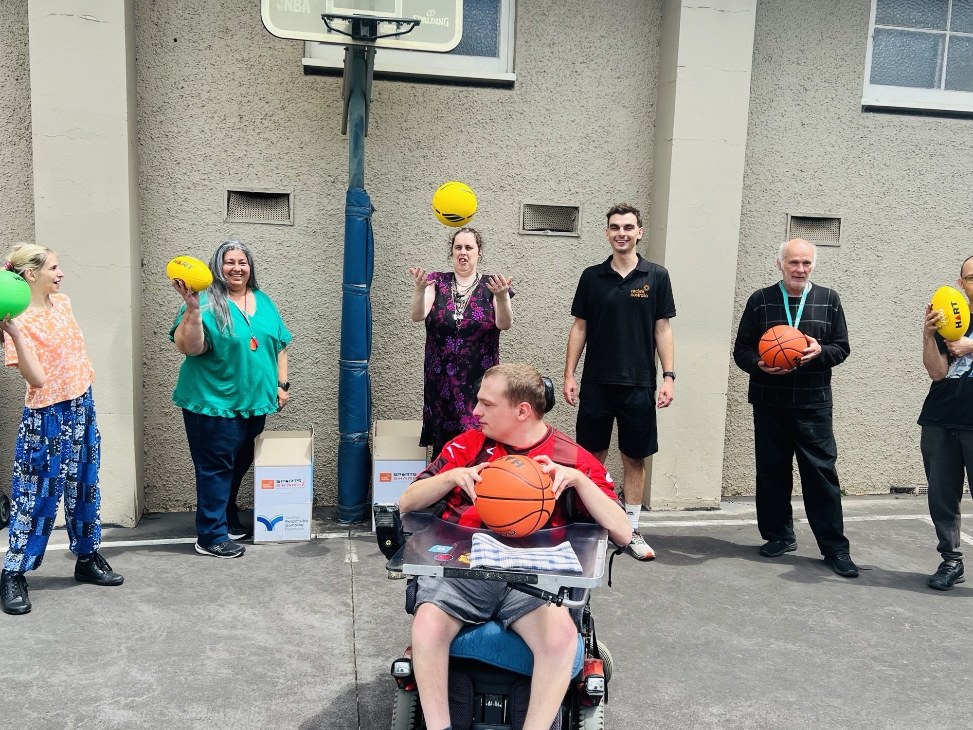 Group of people standing outdoors, holding various sports balls including basketballs, yellow balls, and a green ball, with a person in a motorized wheelchair in front, all smiling or engaging in a casual gathering.