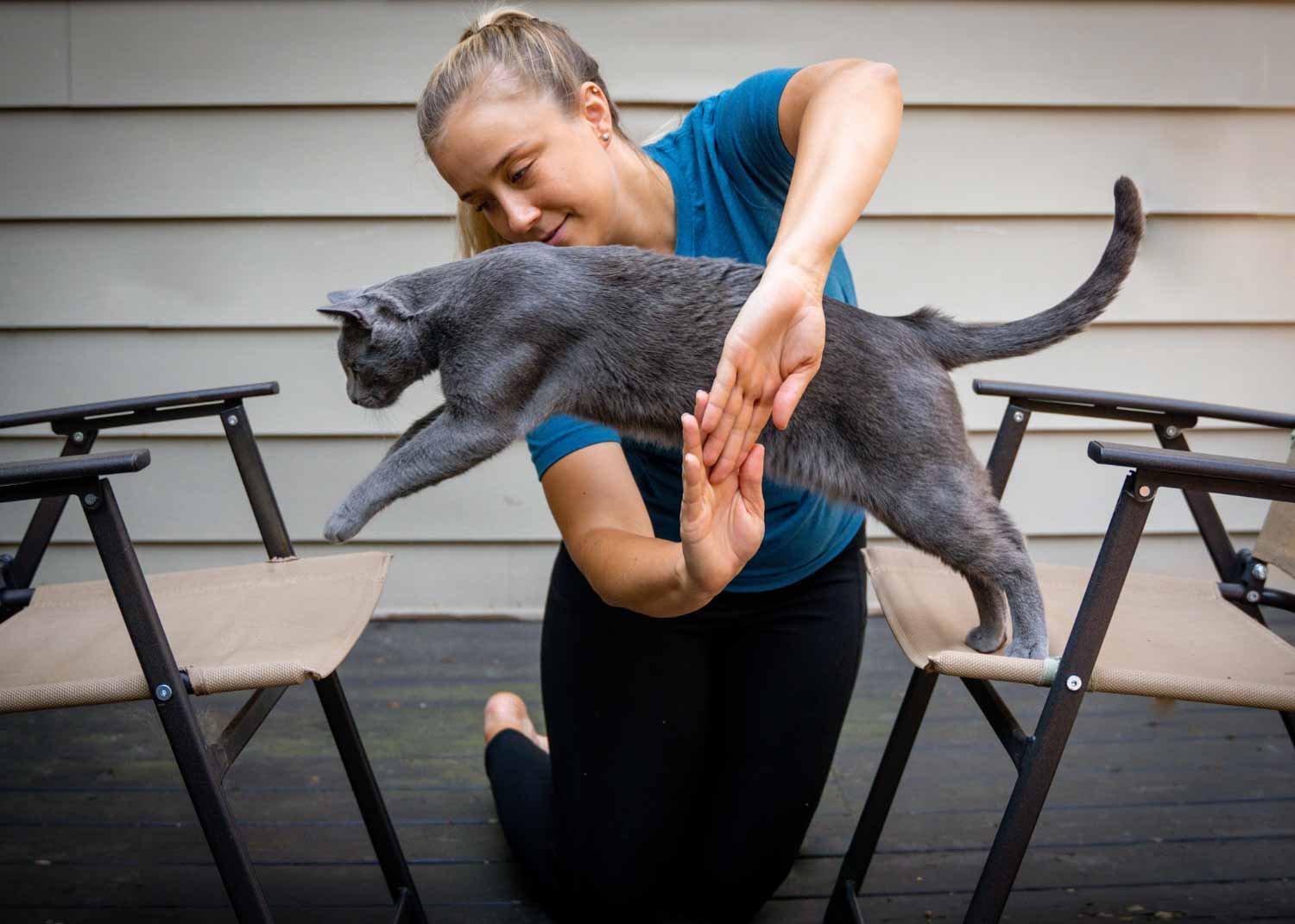 A woman practicing yoga outdoors with a gray cat balanced on a table, as the woman holds the cat's paw and tail.