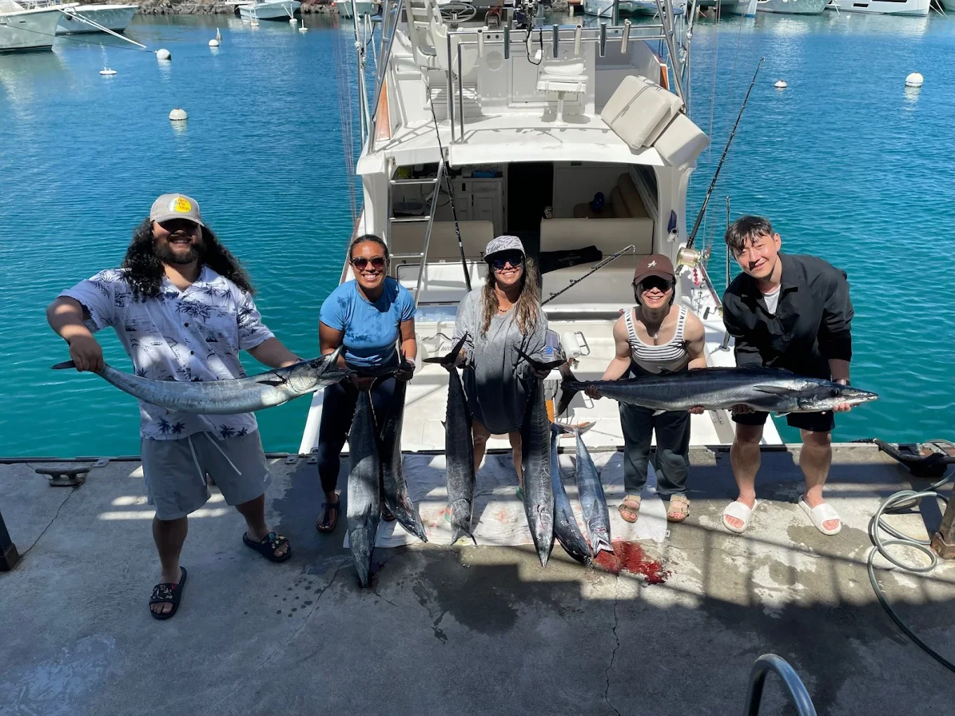 Group of five people standing on a dock, holding large fish they caught, with a boat and marina in the background.
