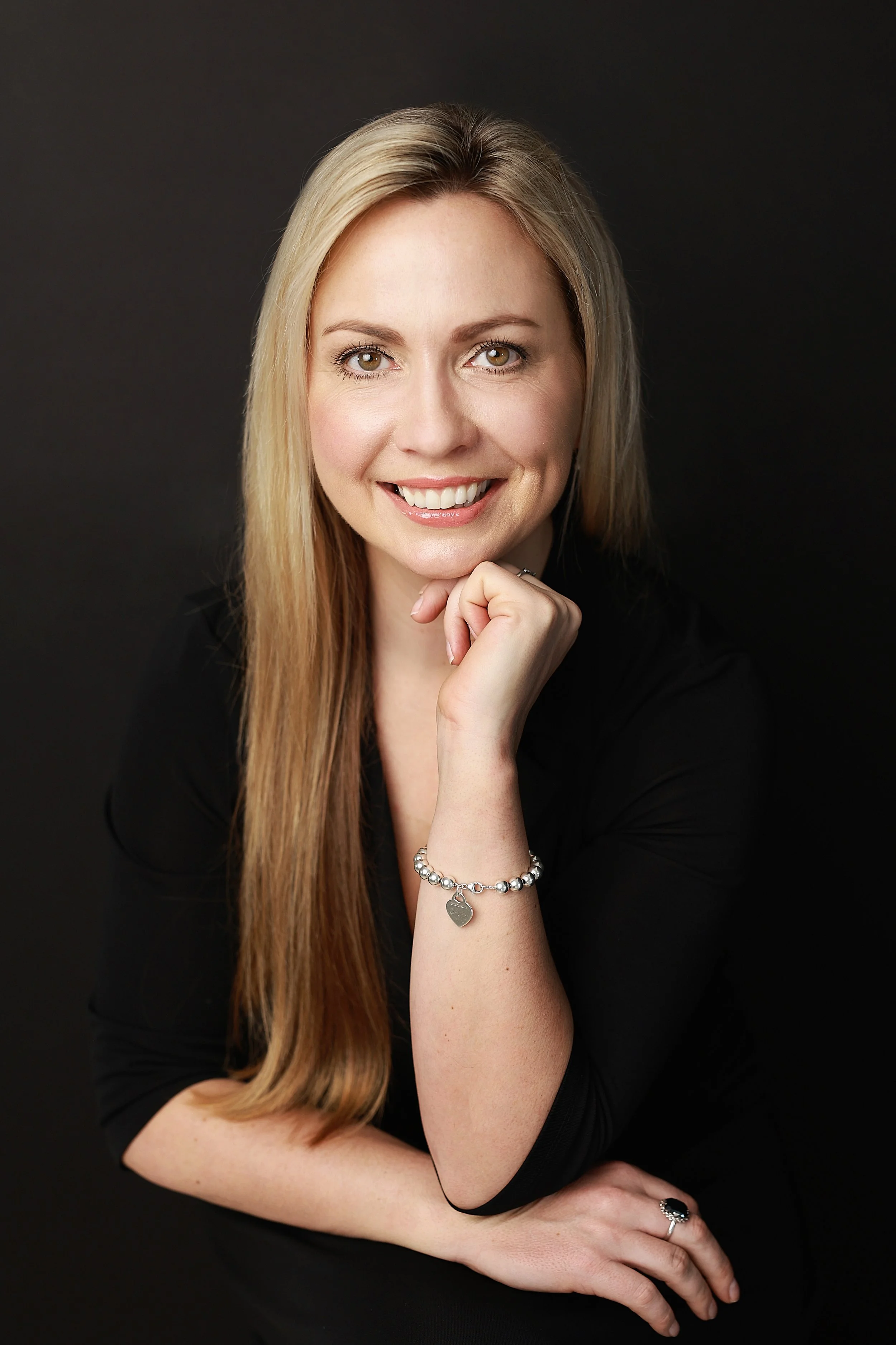 A woman with long blonde hair smiling, wearing a black top, jewelry, and sitting against a dark background.