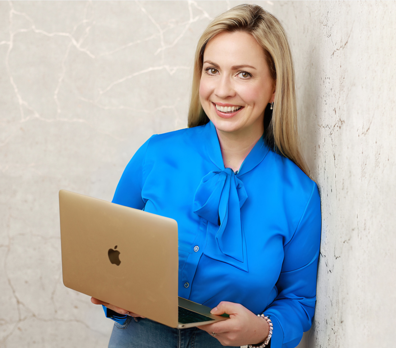A smiling woman with blonde hair wearing a blue blouse with a bow, holding a gold MacBook, standing against a textured white wall.