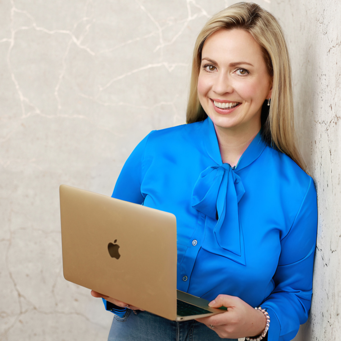 Woman in a bright blue blouse holding a gold MacBook, smiling, leaning against a cracked concrete wall.