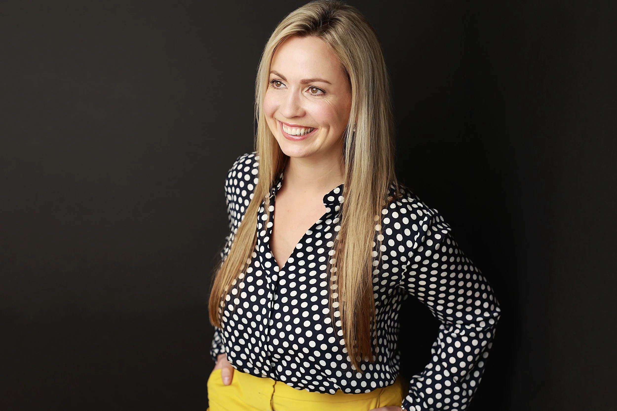A woman with long blonde hair and blue eyes, smiling, wearing a black and white polka dot blouse and yellow pants, standing against a dark background.