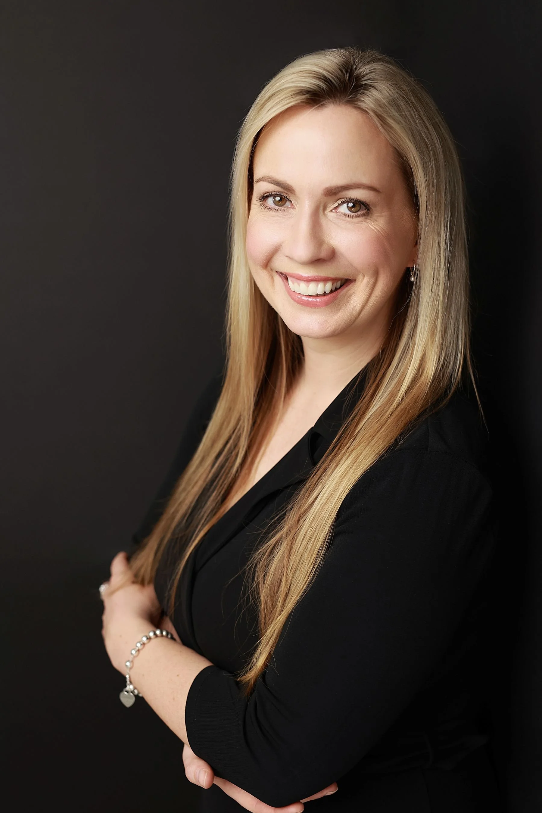 Portrait of a smiling woman with long blonde hair, wearing a black blazer and standing against a dark background.