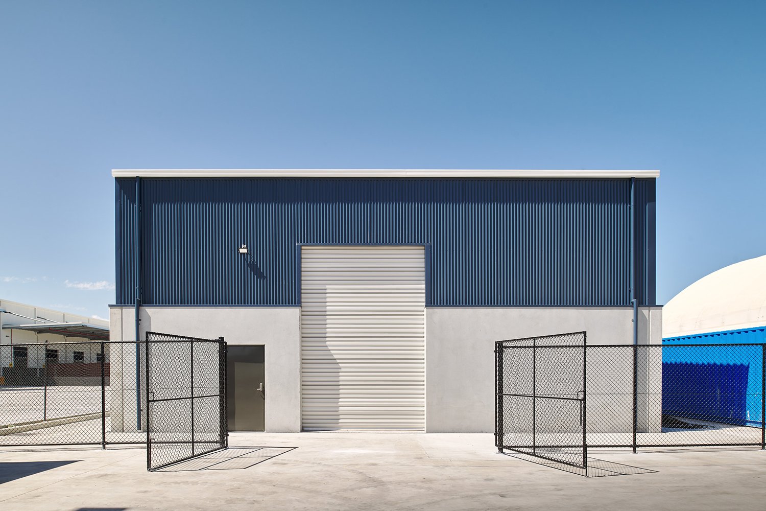 Industrial building with corrugated blue facade, metal roller door, and chain-link fence, under clear blue sky.