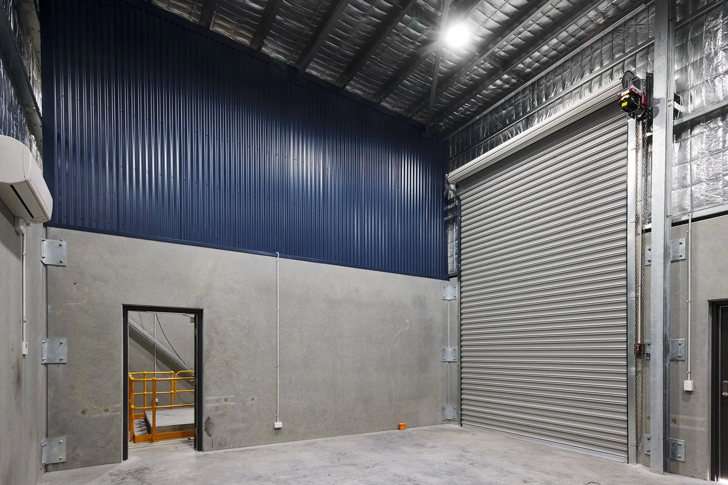 Industrial warehouse interior with corrugated metal walls and ceiling, concrete floor, and a large closed roller shutter door.