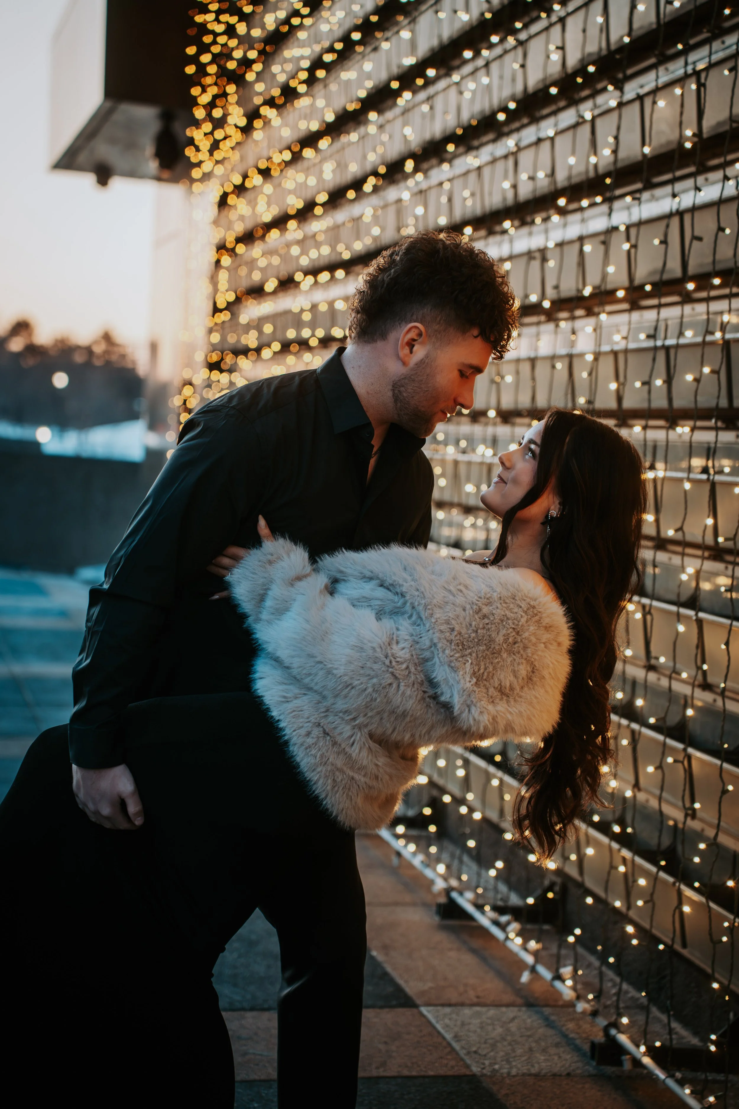 A couple sharing an intimate moment in front of a wall decorated with warm holiday string lights at sunset.