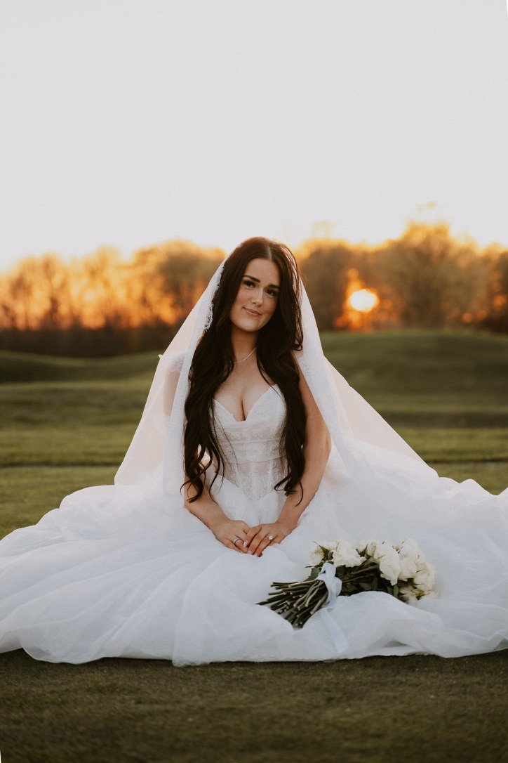 A woman in a wedding dress sitting on grass with a bouquet of white roses in front of her. The background shows a sunset with trees.