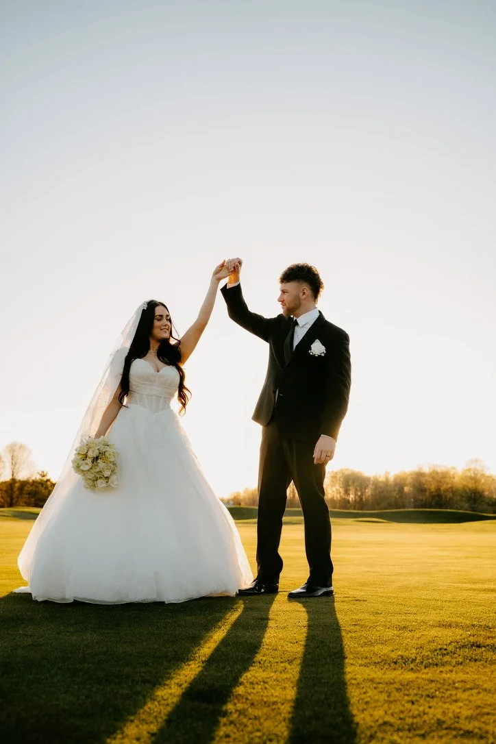 Bride and groom dancing on a golf course at sunset, with the bride in a white wedding gown holding a bouquet and the groom in a black suit, they are smiling and holding hands.