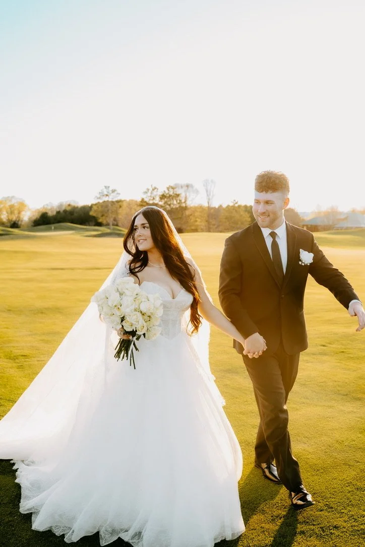 A bride and groom holding hands, walking on a green field during sunset, with the bride in a white wedding dress holding a bouquet of white roses and the groom in a black tuxedo.