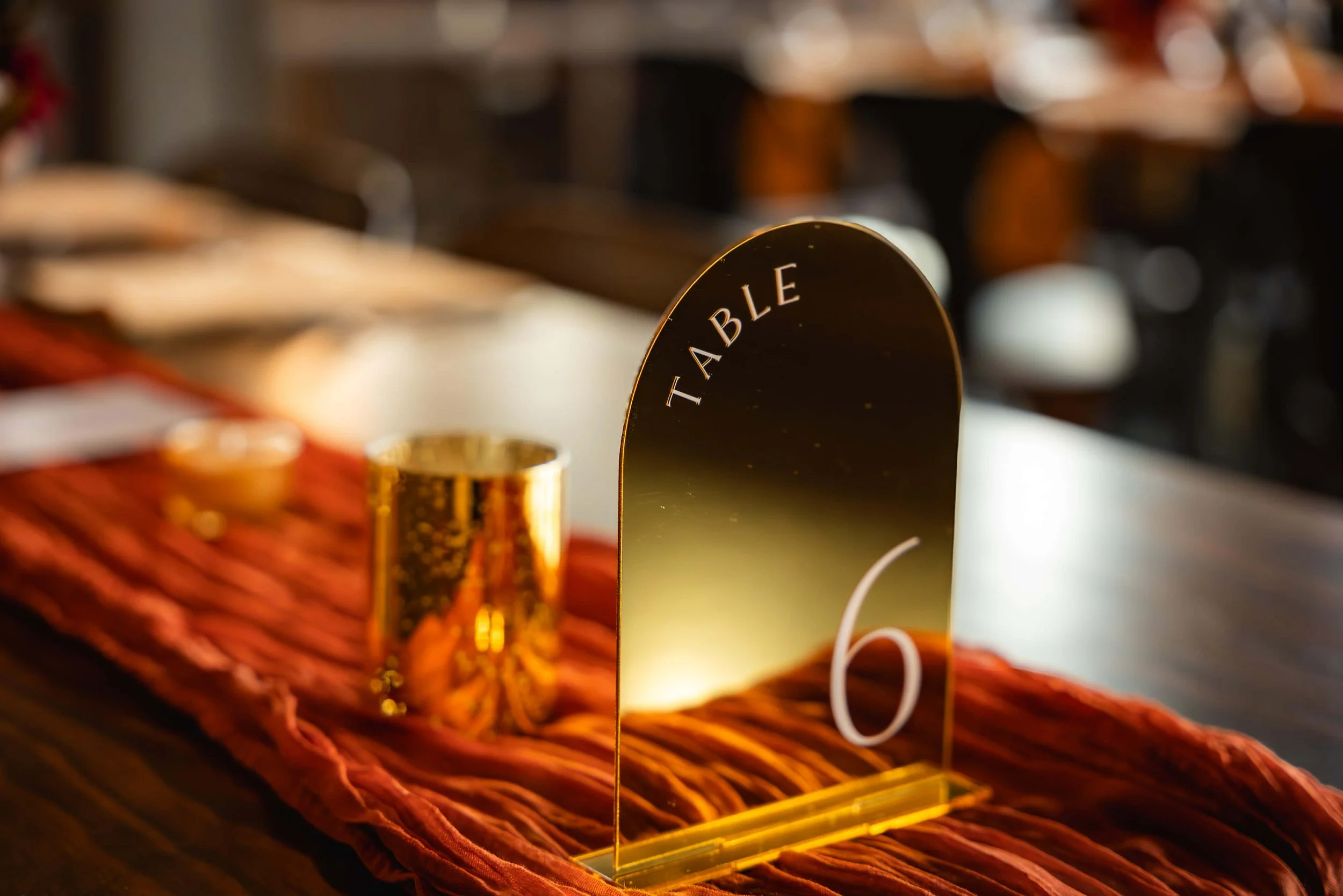 Table number sign with the number 6 and the word 'TABLE' on a gold stand, placed on a red table runner with a small gold metallic cup next to it.