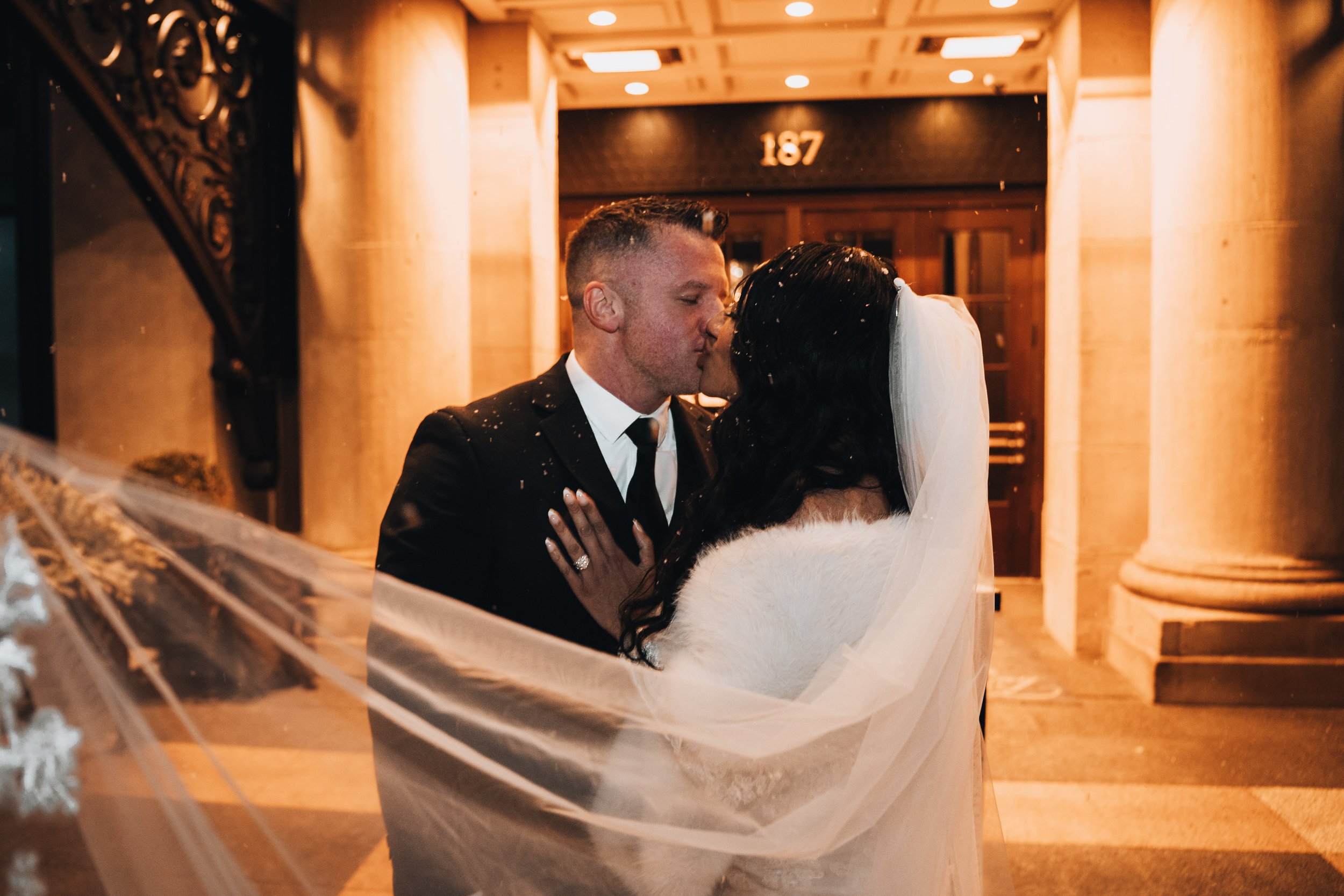 A newlywed couple sharing a kiss outside a building, with the groom in a black suit and the bride in a white wedding dress and veil, surrounded by falling confetti.