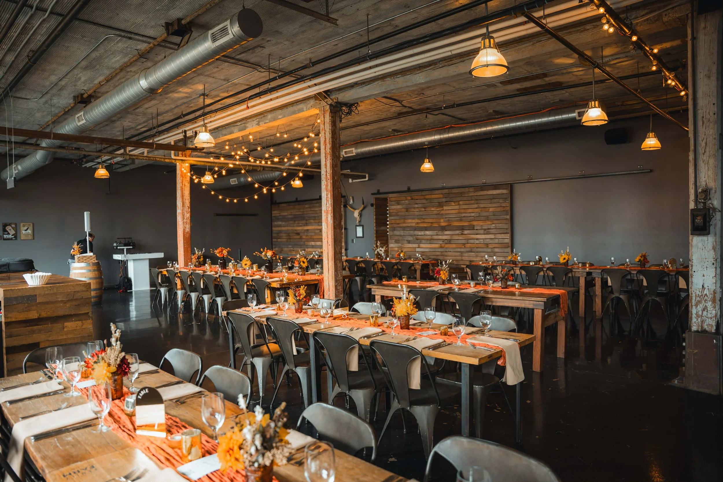 Interior of a rustic event space set for a celebration with long wooden tables decorated with floral centerpieces, wine glasses, and napkins, featuring string lights hanging from the ceiling and a wooden wall backdrop.