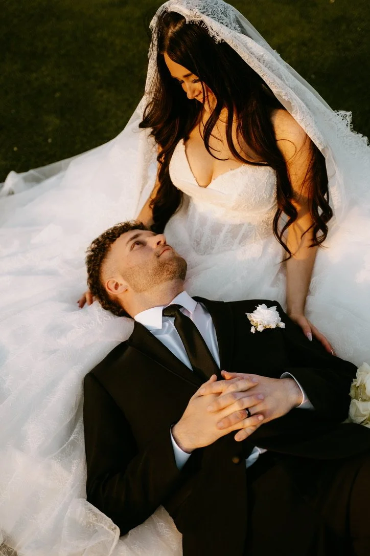 Bride and groom on their wedding day, with the bride sitting and the groom lying on the ground, looking up at her.