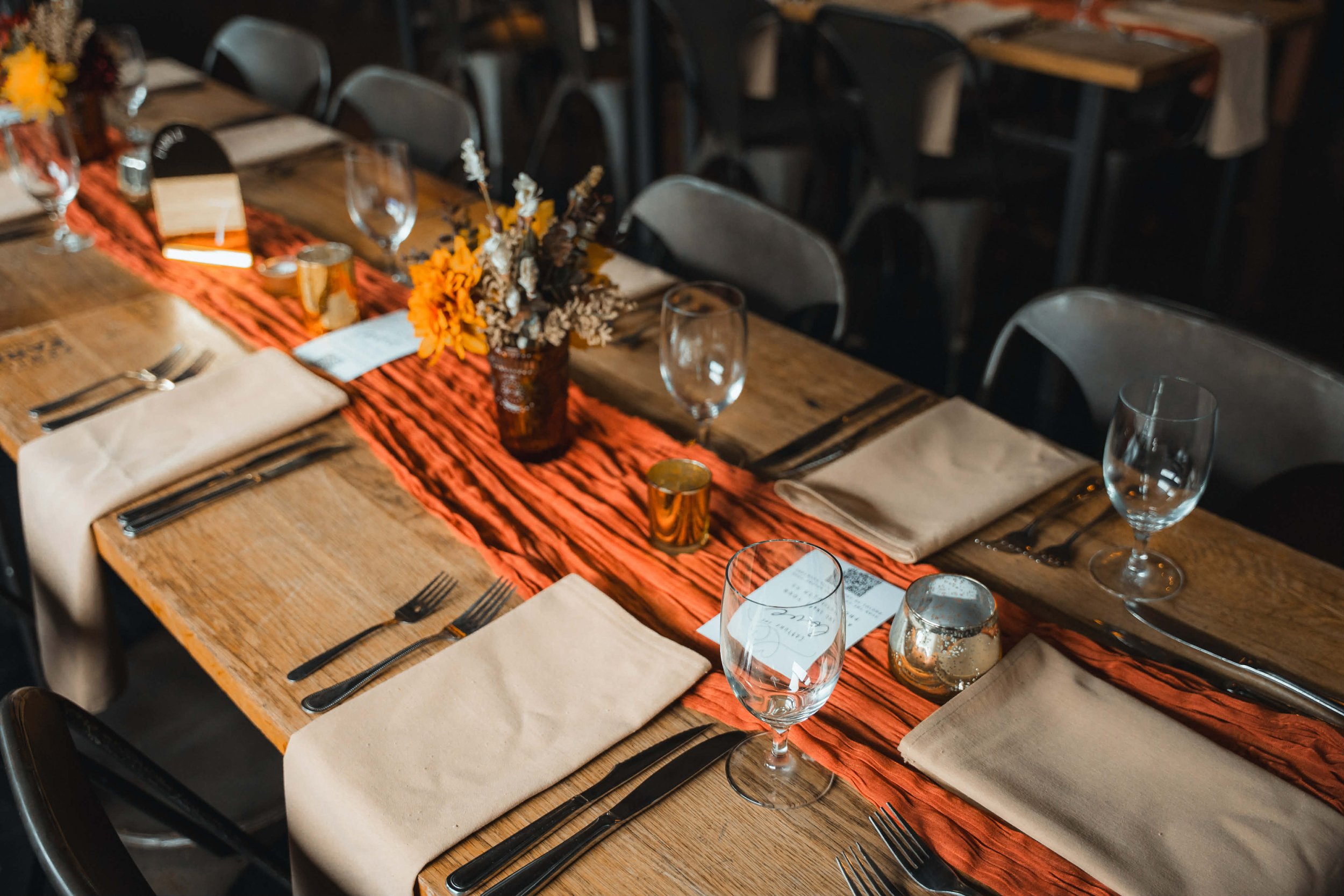A dining table set for a formal event features beige napkins, silverware, empty wine glasses, and small decorative candles. A rust-colored table runner runs down the center, topped with a flower arrangement in a brown vase and decorative candles.