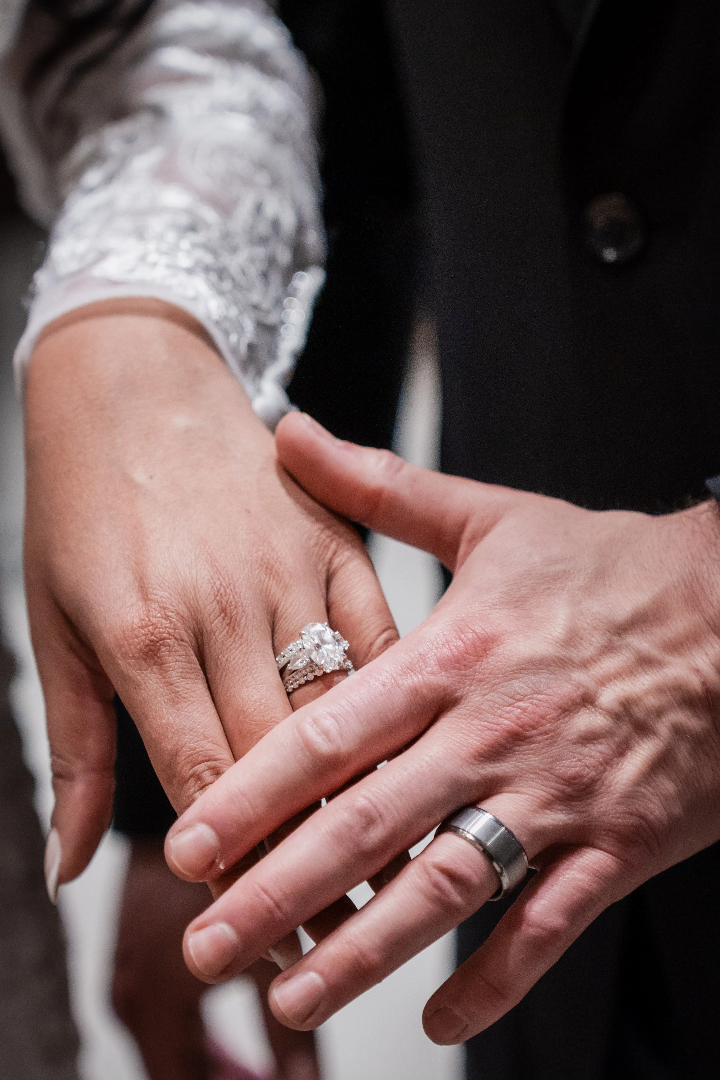 Close-up of a bride and groom's hands showing wedding rings; the bride's hand has an intricate engagement ring and wedding band, the groom's hand has a simple band.