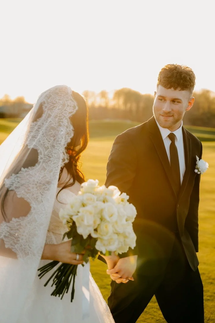 A bride and groom holding hands during a wedding photoshoot outdoors at sunset, with the bride holding a bouquet of white roses, and the bride wearing a lace veil and dress, while the groom is dressed in a black suit and tie.