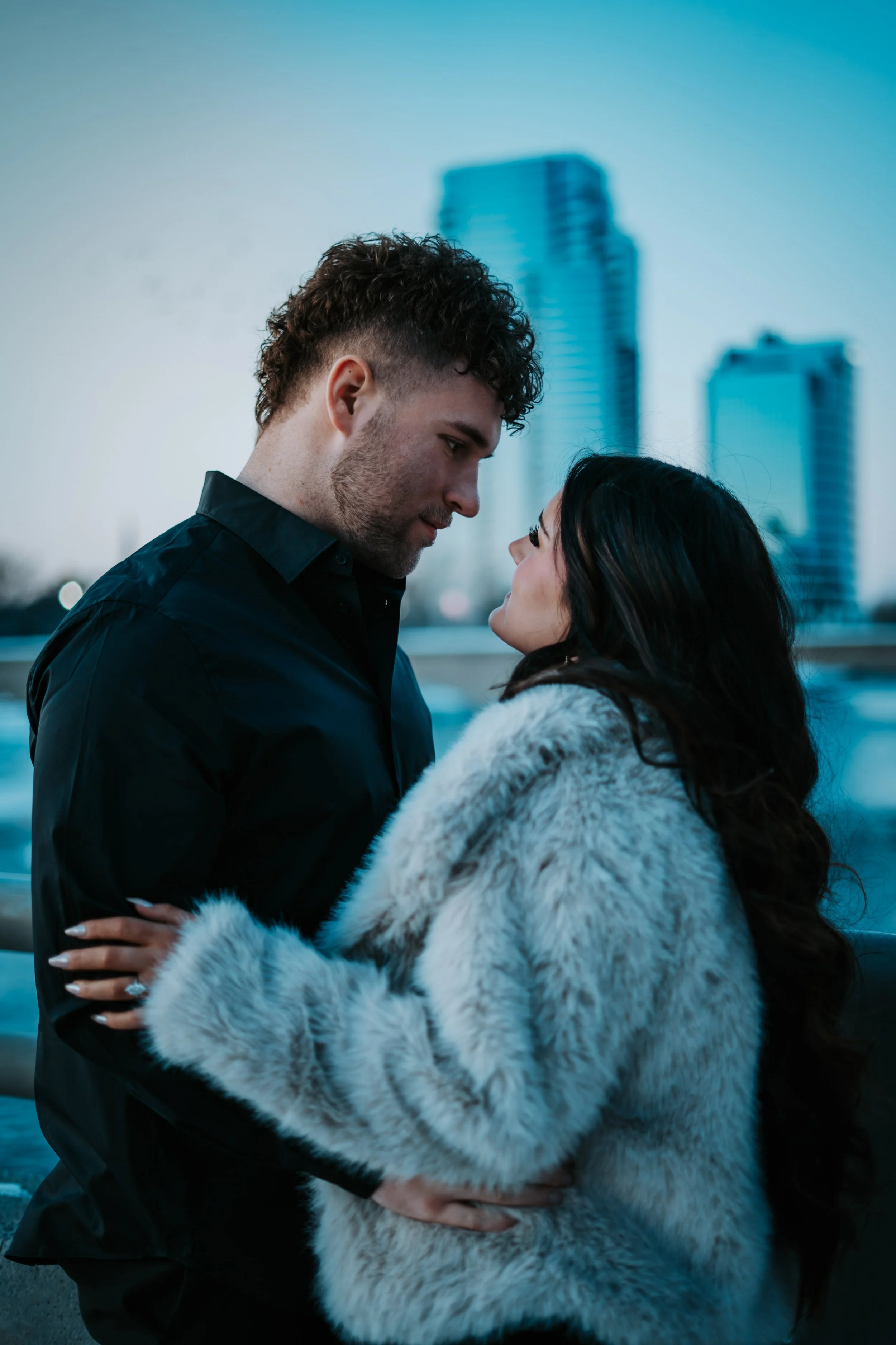 A couple standing close together outdoors with city buildings in the background, gazing into each other's eyes.