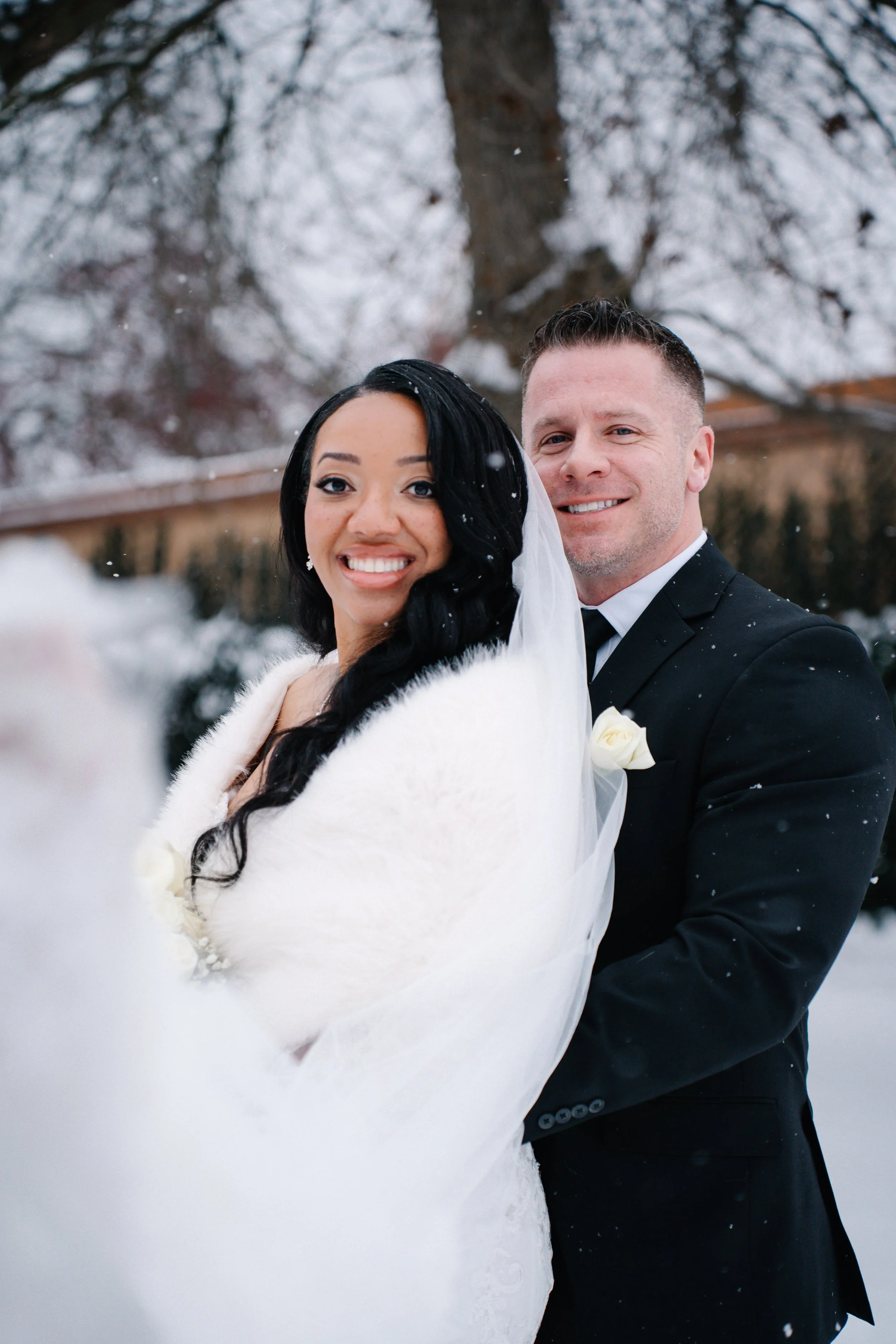 A bride and groom on their wedding day, standing outdoors in a snowy setting with trees in the background. The bride has long black hair and is wearing a white fur stole and a veil, and the groom is in a black suit with a white shirt and a white bout