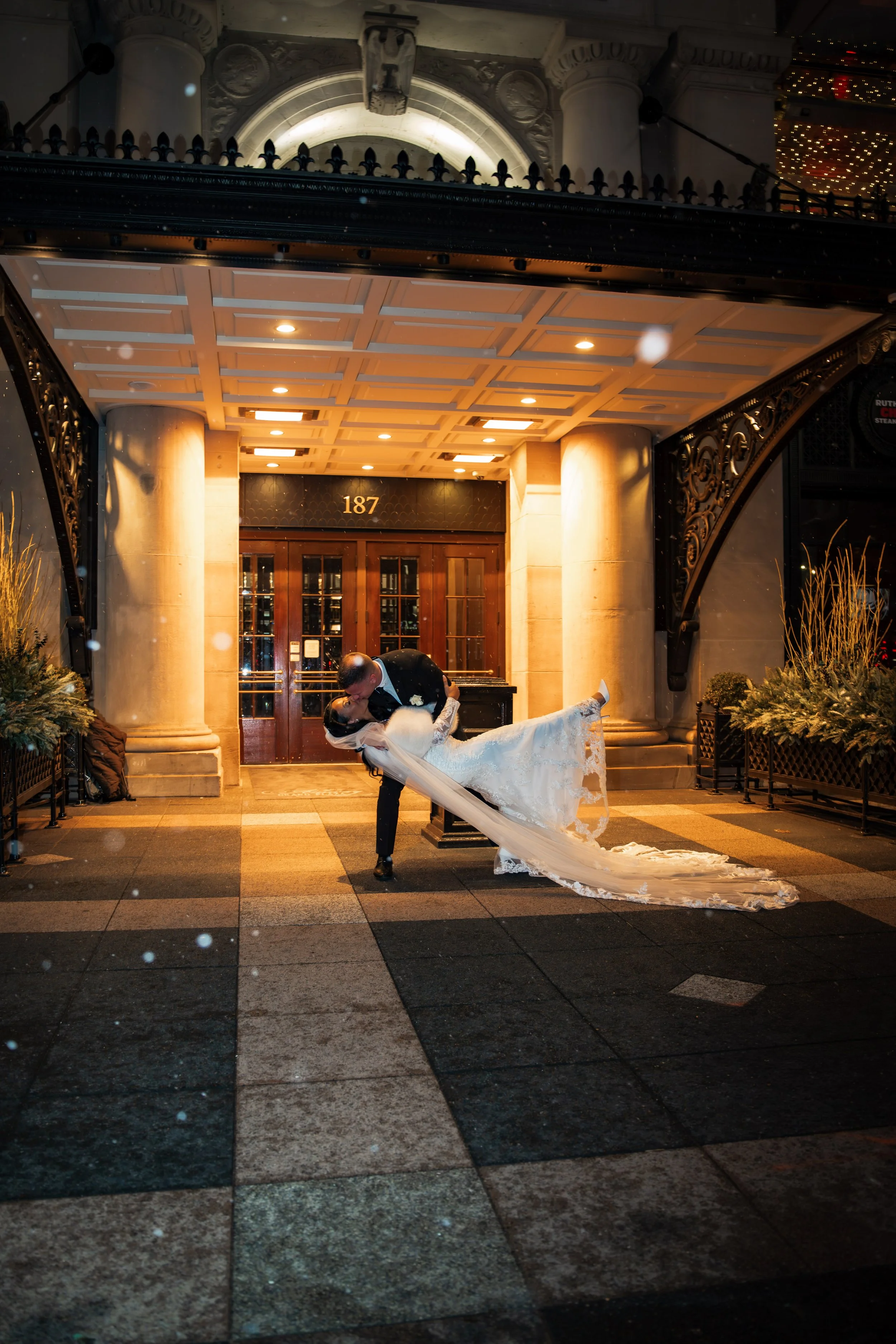 A newlywed couple sharing a kiss outside a building at night, with the groom dipping the bride. The building has the number 187 above the entrance and is decorated with columns and ornate ironwork. The bride is in a white wedding dress with a long tr