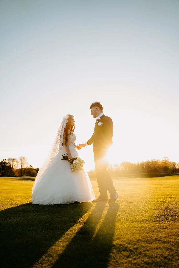 A bride and groom holding hands on a grassy field during sunset, dressed in wedding attire, with sunlight creating a warm glow.