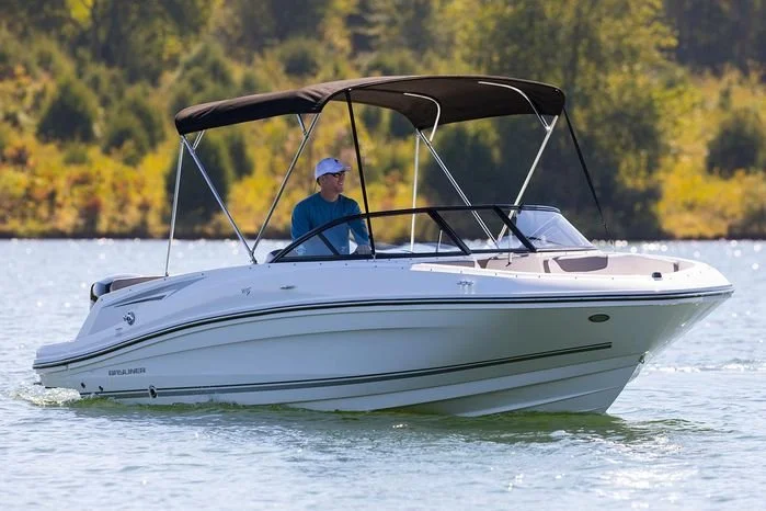 Man in a blue shirt and white hat boating on a white motorboat with a black canopy on a lake, with trees in the background.