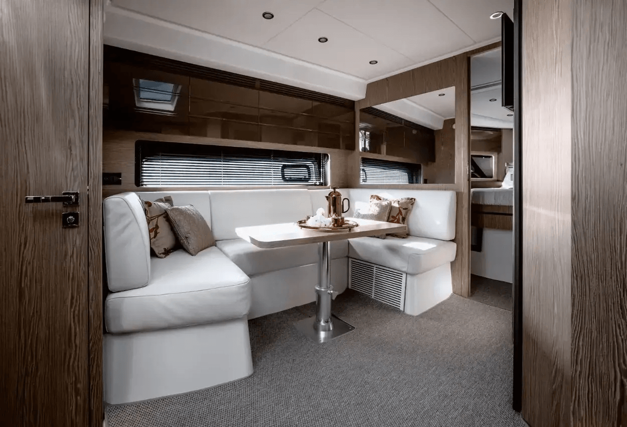 Interior of a modern yacht with a cozy dining area featuring white cushioned seating, a wooden table, decorative pillows, and a French press coffee set. The design includes wood paneling, carpet flooring, and large windows with blinds.