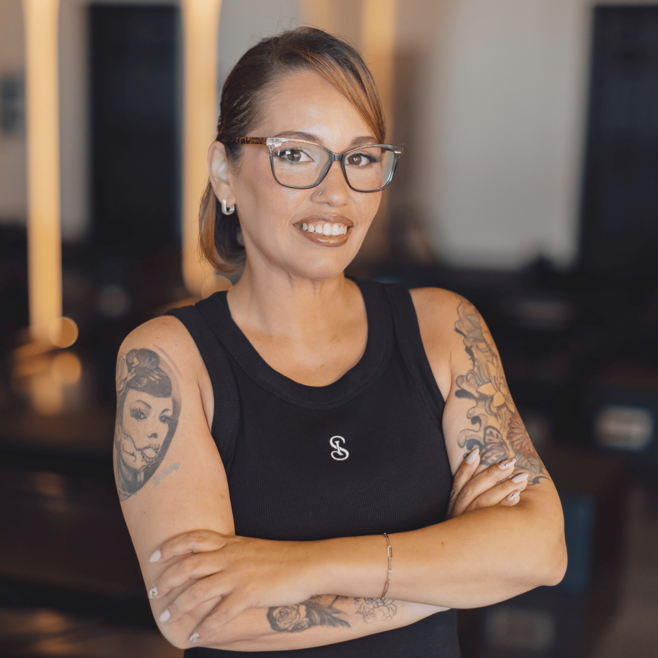 A woman with tattoos on her arms and glasses smiling, crossed arms, wearing a black tank top, in an indoor setting with blurred background.