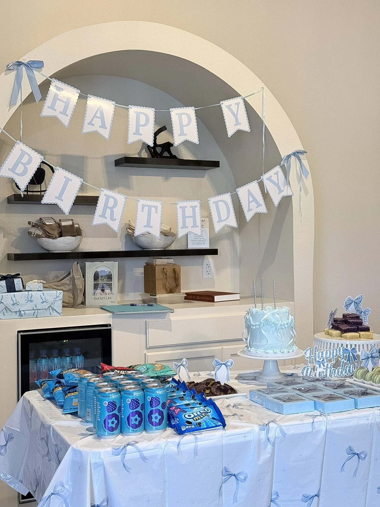 Party dessert table decorated with blue and white theme, featuring a "Happy Birthday" banner, a white wedding cake with blue ribbons, various snacks, and a collection of blue cans of flavored sparkling water.