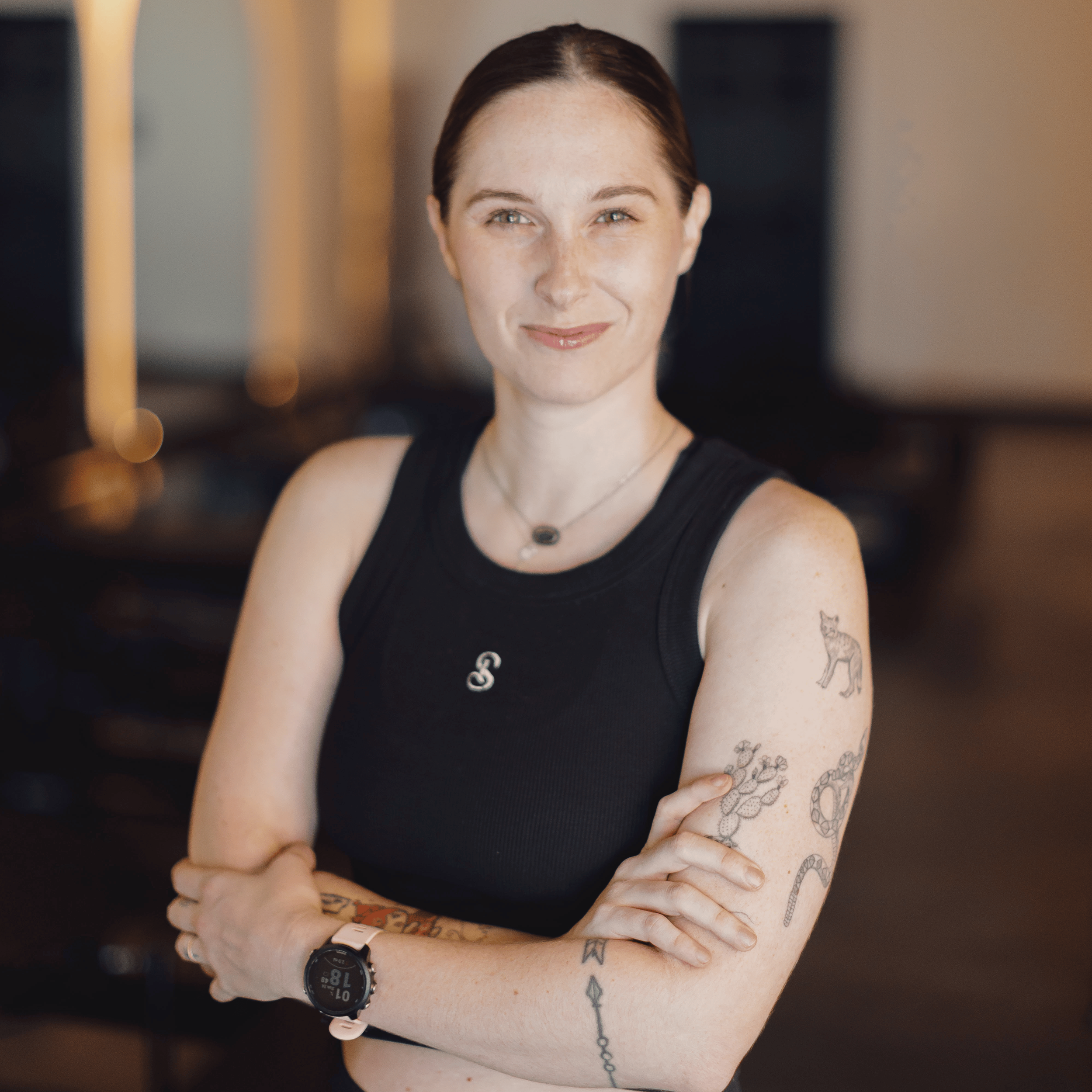 Close-up of a woman with short dark hair, wearing a black sleeveless top, showing colorful tattoos on her arms and chest, smiling and standing with crossed arms in a warmly lit indoor setting.