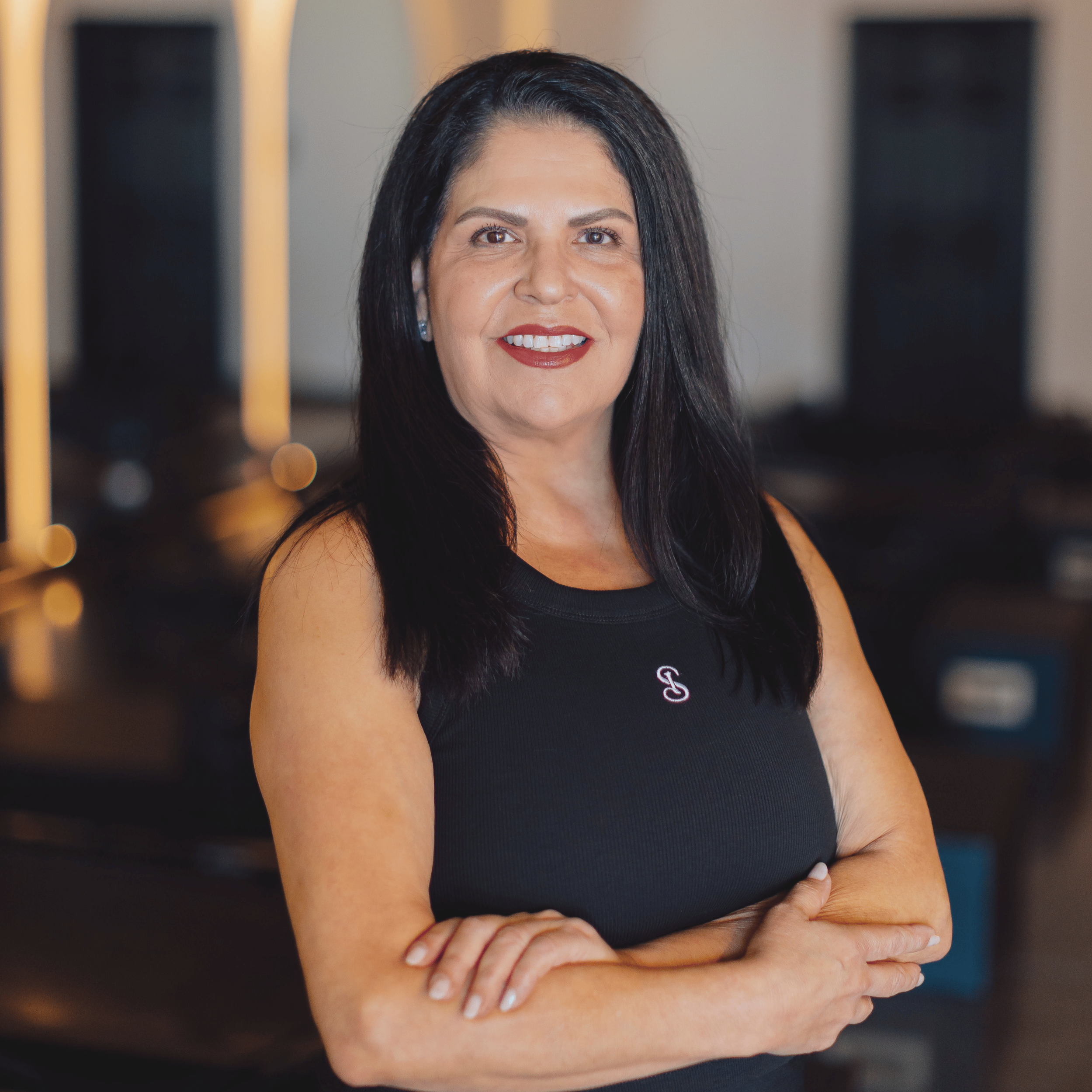 Woman with long black hair smiling, wearing a black top with a small embroidered logo, standing indoors with blurred background.