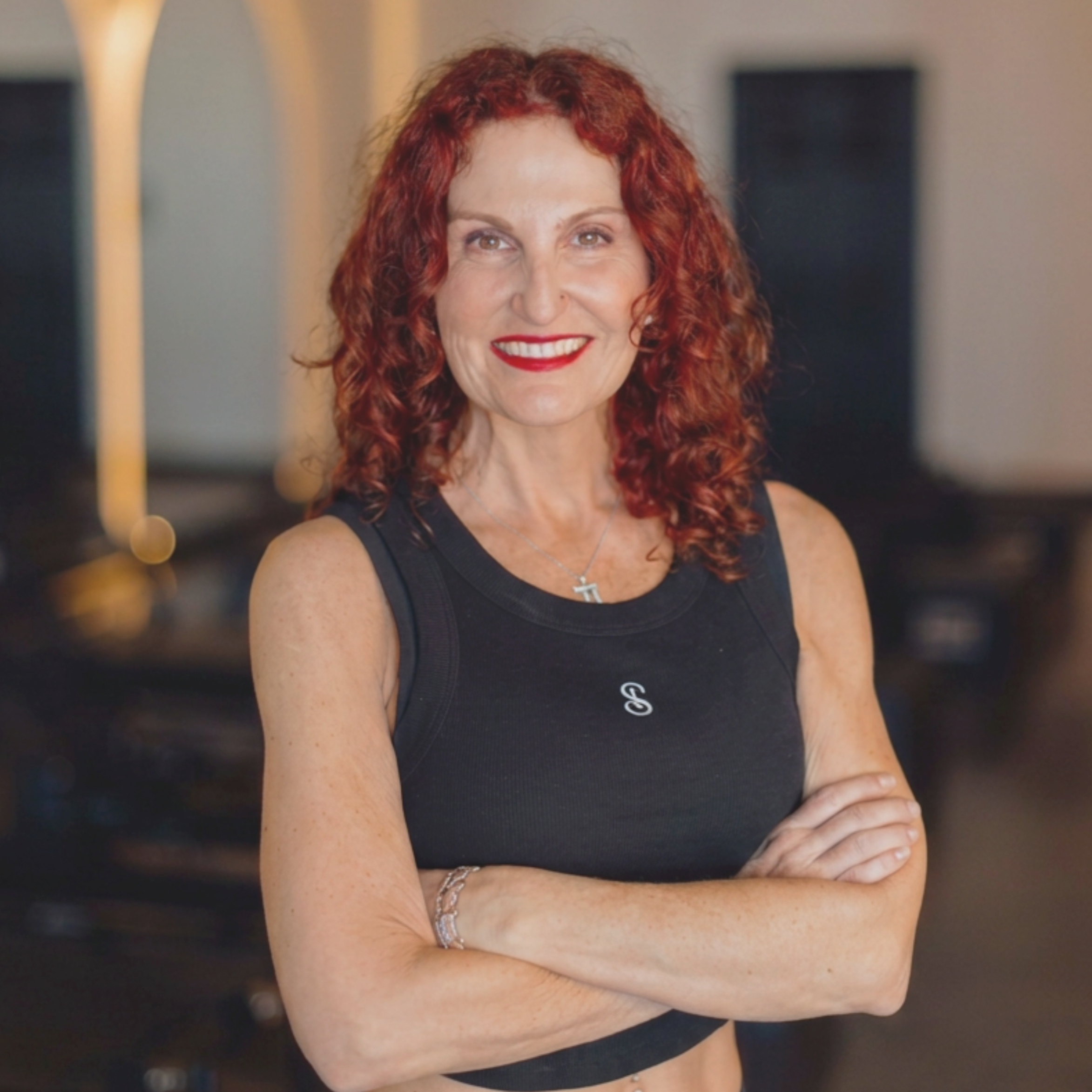 A woman with curly red hair wearing a black sleeveless top, smiling with her arms crossed, in an indoor setting.