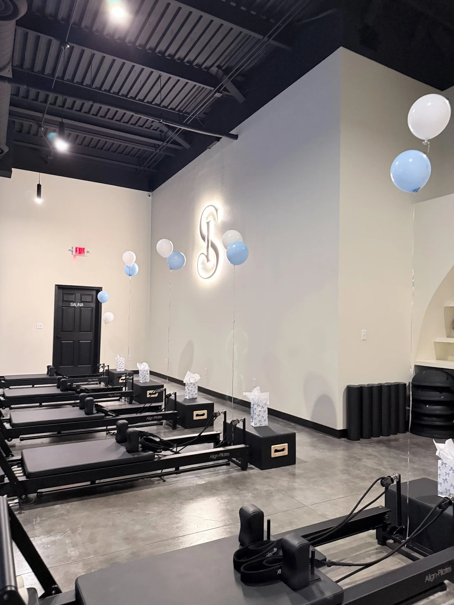 Interior of a fitness studio with Pilates reformer machines lined up on a polished concrete floor. Light balloons hang from the ceiling and tissue boxes are placed on the floor, indicating a celebration or special event.
