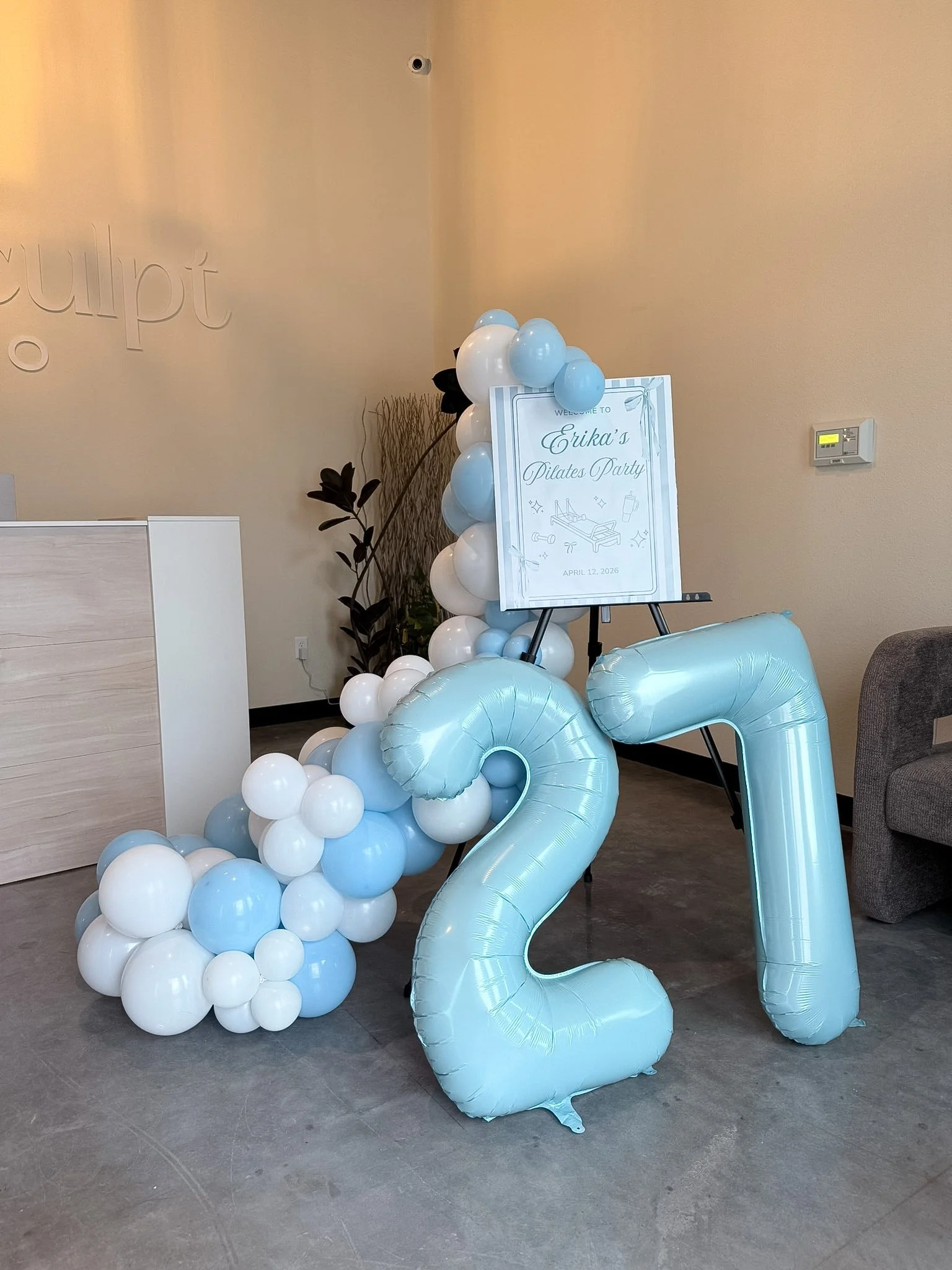 Blue and white balloons arranged in a decorative arch with a sign that reads "Erika's Pilates Party" and the date April 12, 2026, in front of a beige wall.