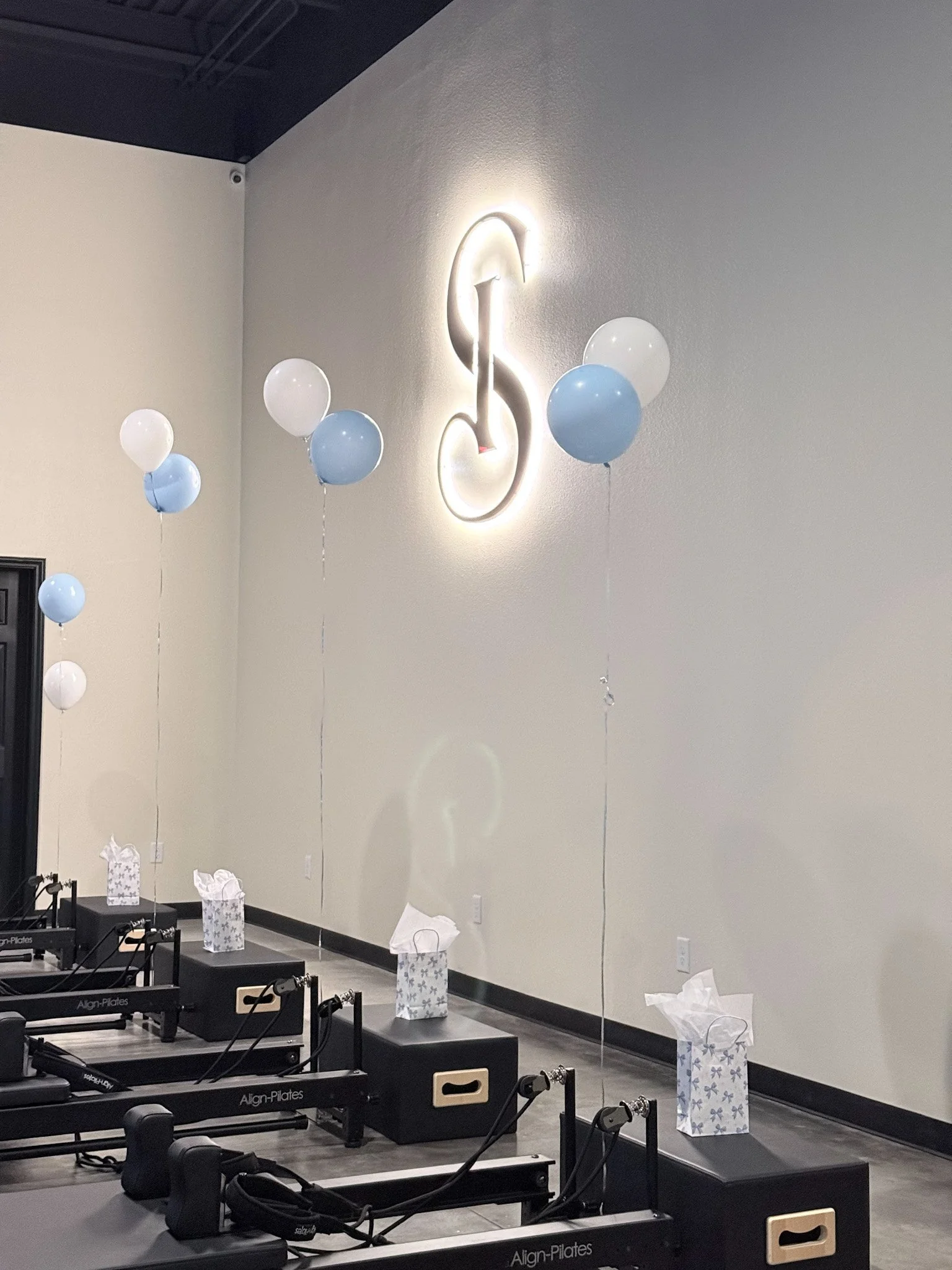 A room with Pilates reformer machines on a black mat, white and blue balloons attached to tall paper gift bags, and a wall decorated with a glowing treble clef musical symbol.
