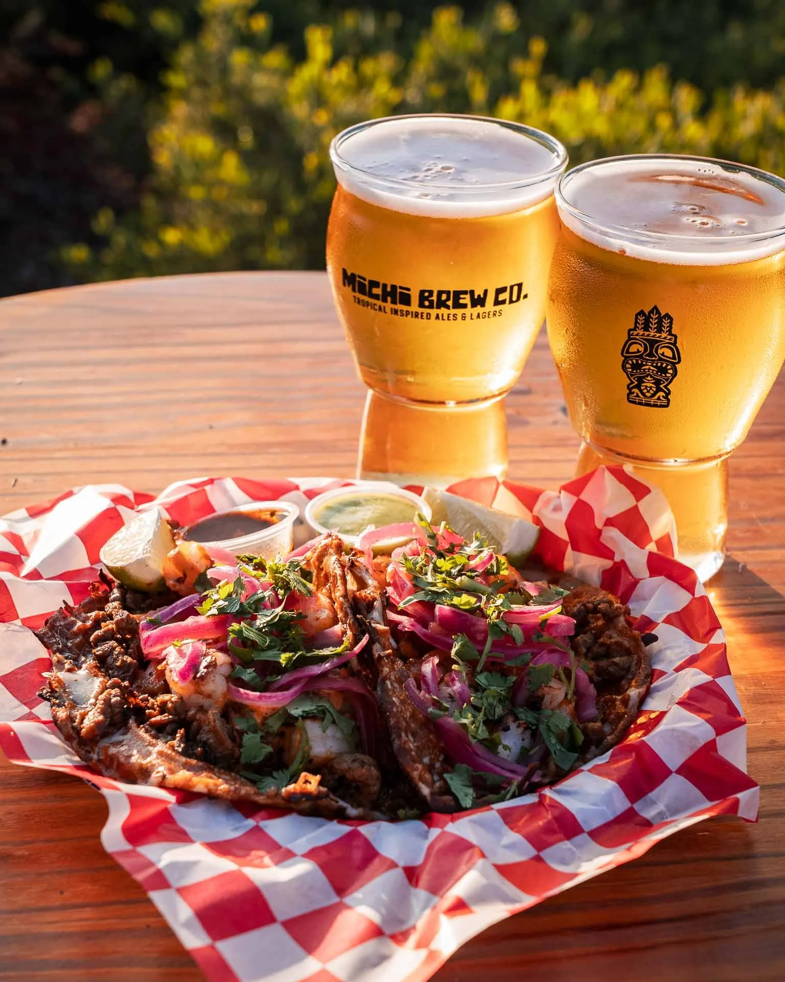 Two glasses of beer with foam, a plate of tacos with meat, onions, cilantro, and lime wedges, and small containers of sauce on a checkered paper in an outdoor setting.