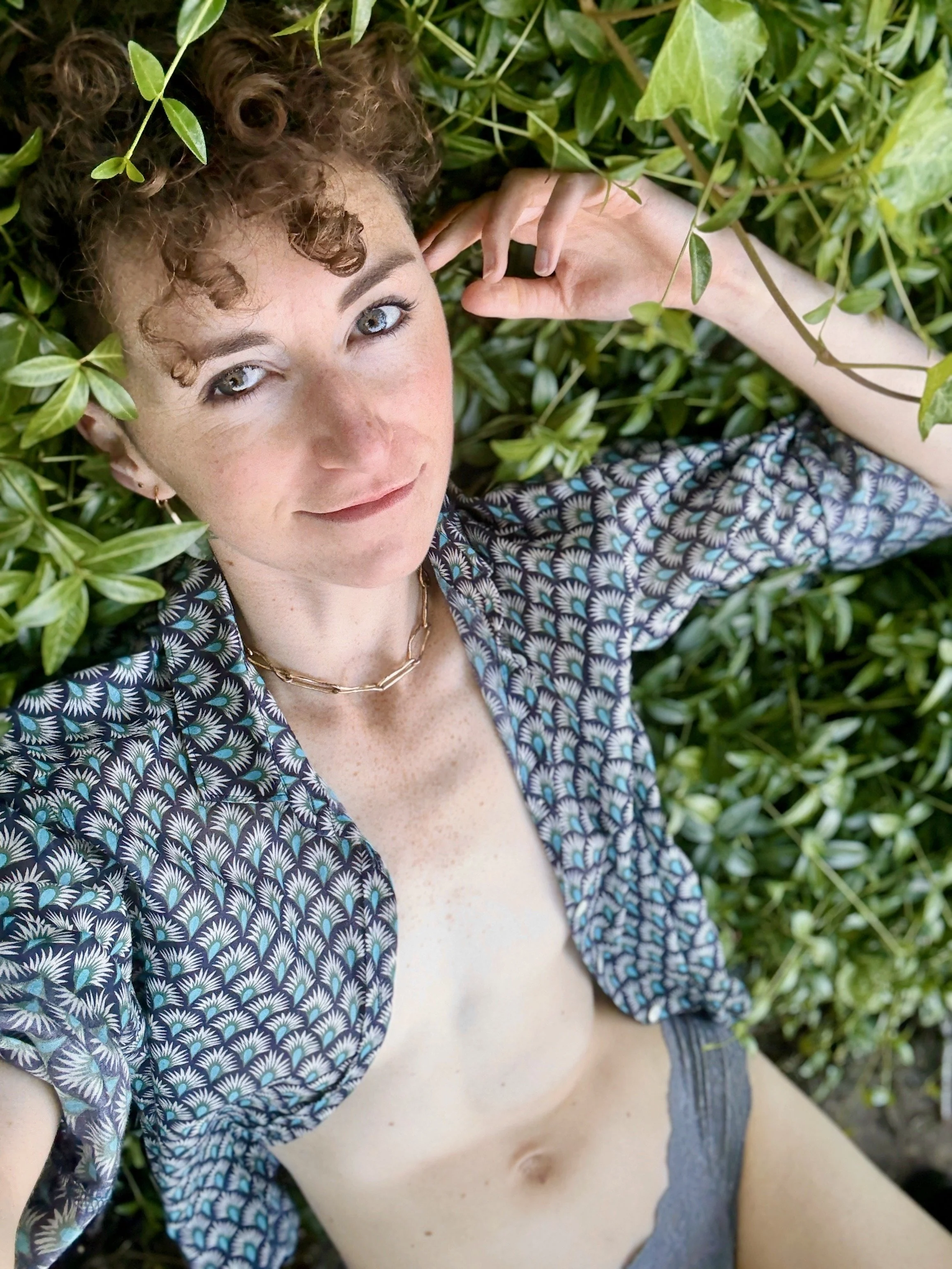 A woman with curly red hair and light eyes lying among green leaves, wearing a patterned shirt, grey underwear, a gold necklace, and a small earring.