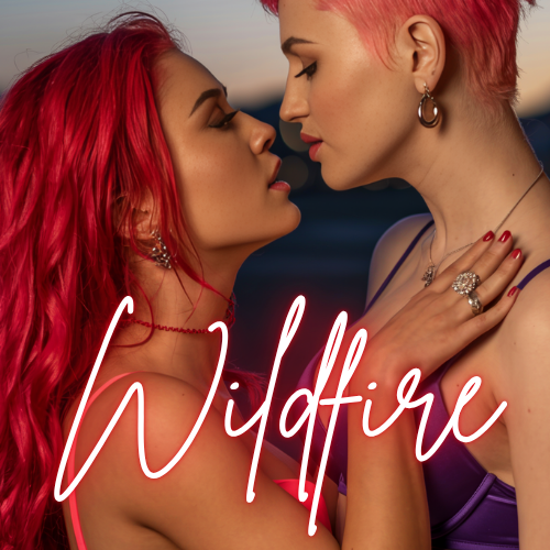 Two women with colorful hair, one with red hair and the other with pink hair, close together at sunset with the word 'Wildfire' written in cursive.