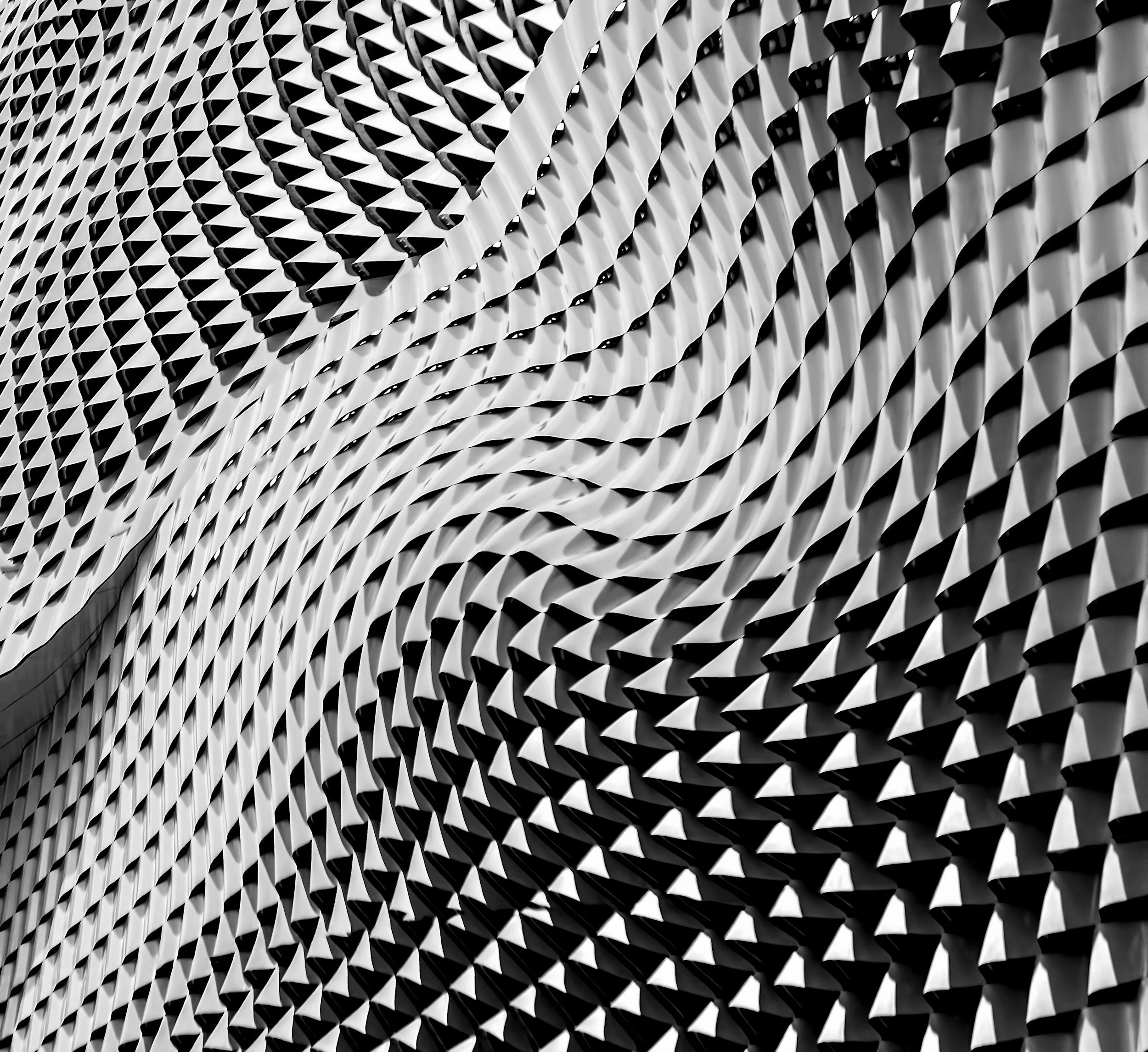 Close-up of a modern building facade with a repeating geometric pattern of diamond-shaped openings, creating a textured and visually dynamic surface in black and white.